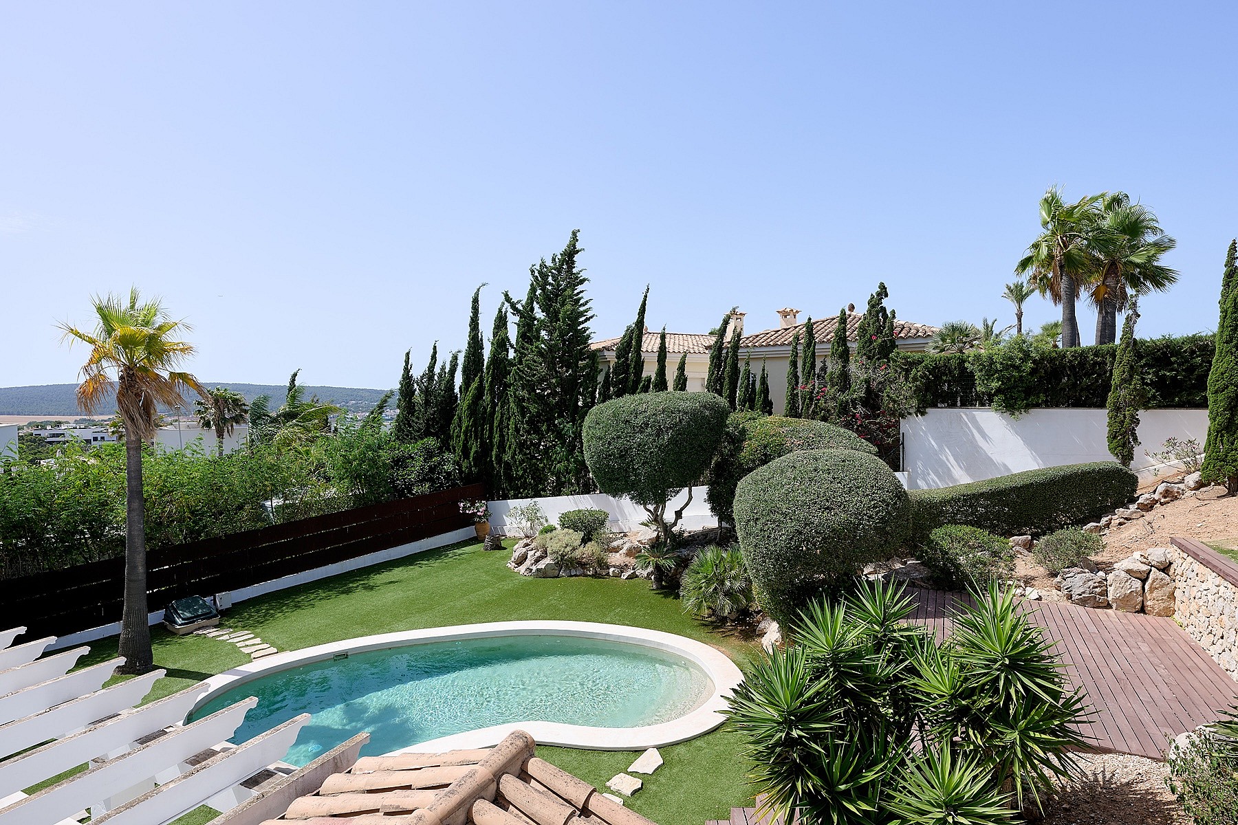Vista desde tarima de madera sobre piscina ovalada, césped artificial, topiaria y palmeras con montañas al fondo