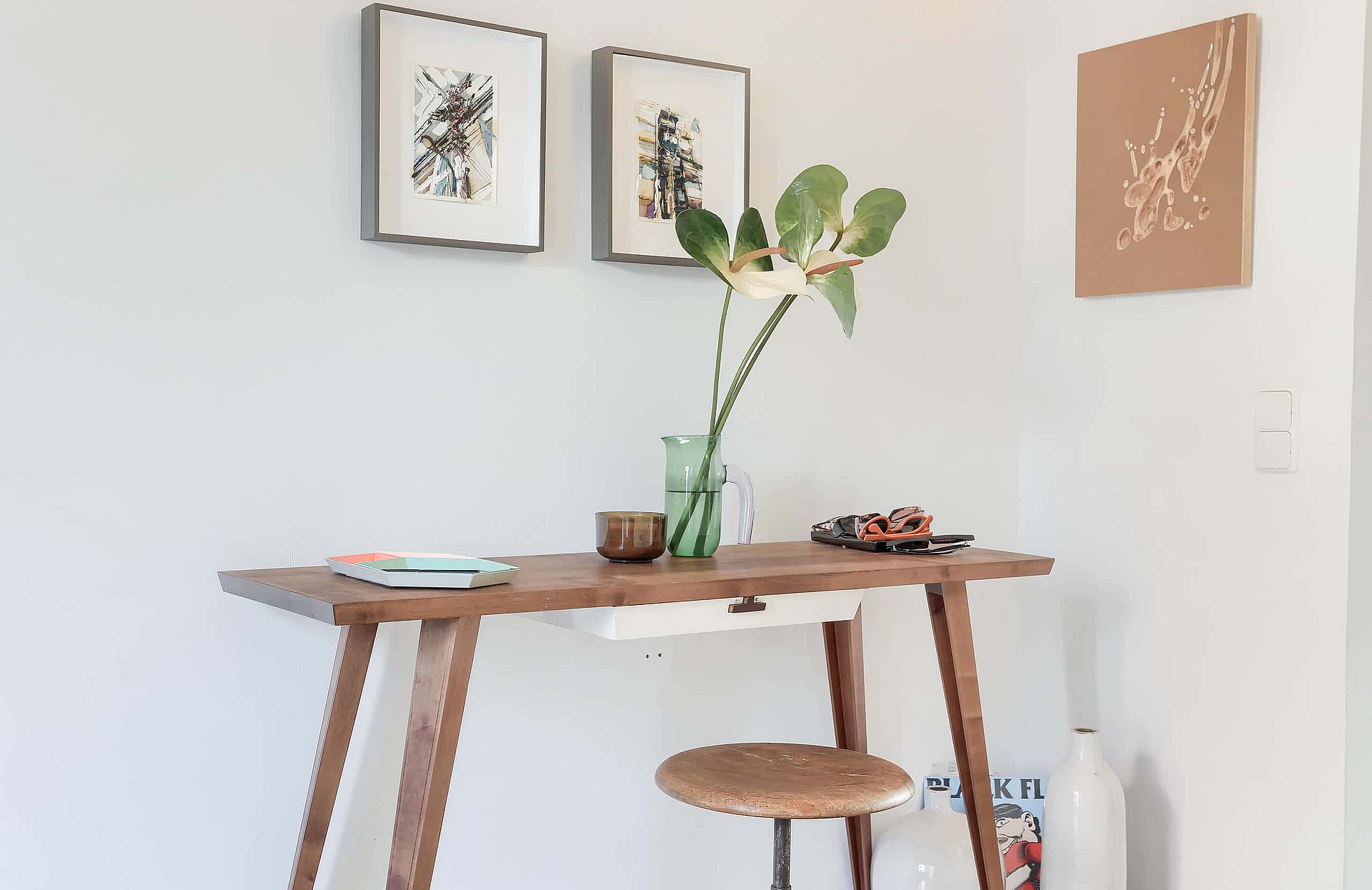 A wooden writing desk with a matching stool against a white wall, featuring framed abstract art and a glass vase with greenery.