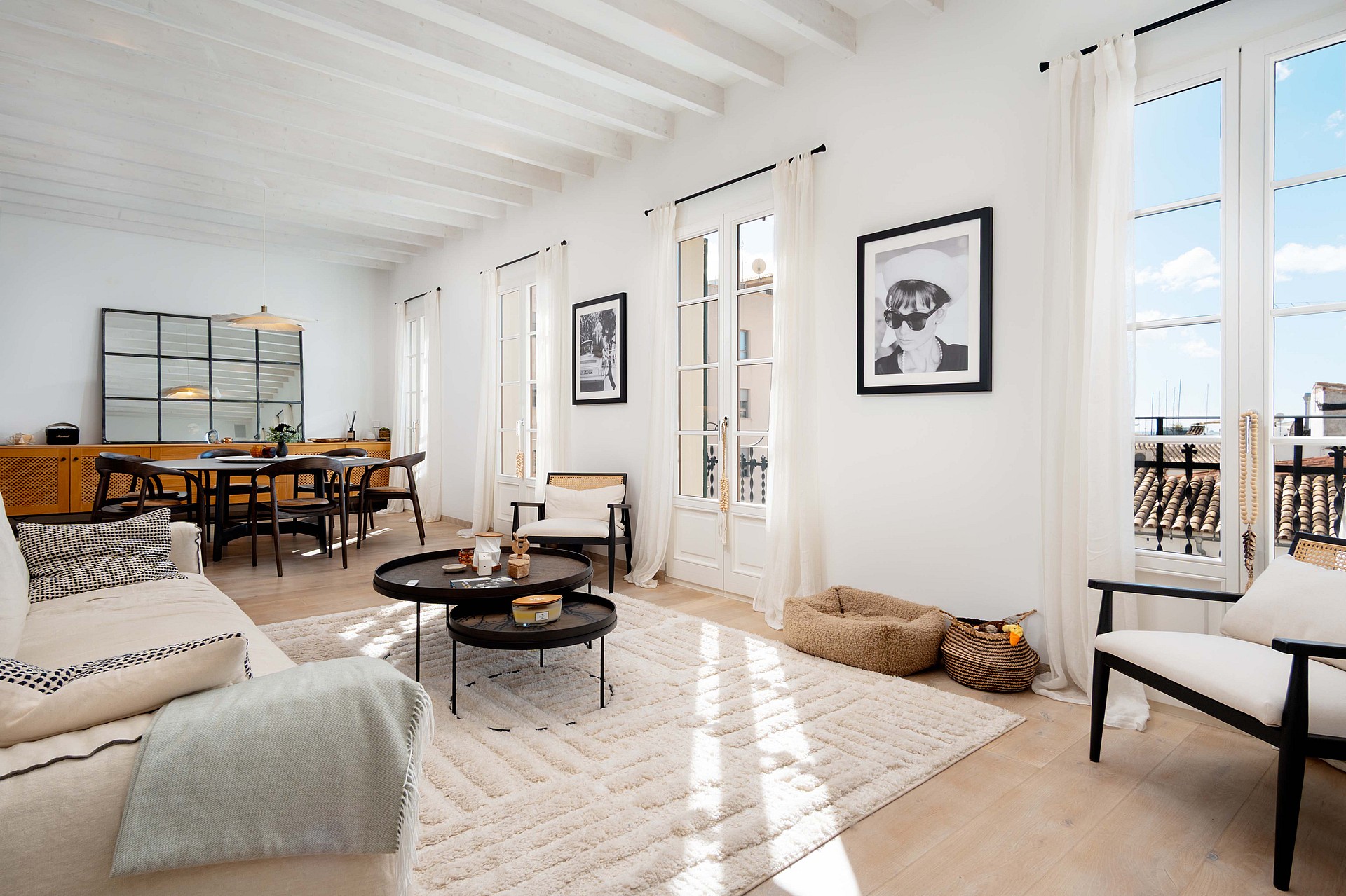 A bright, open-plan living room with white walls, exposed white ceiling beams, a textured area rug, and three sets of French doors leading to a balcony.