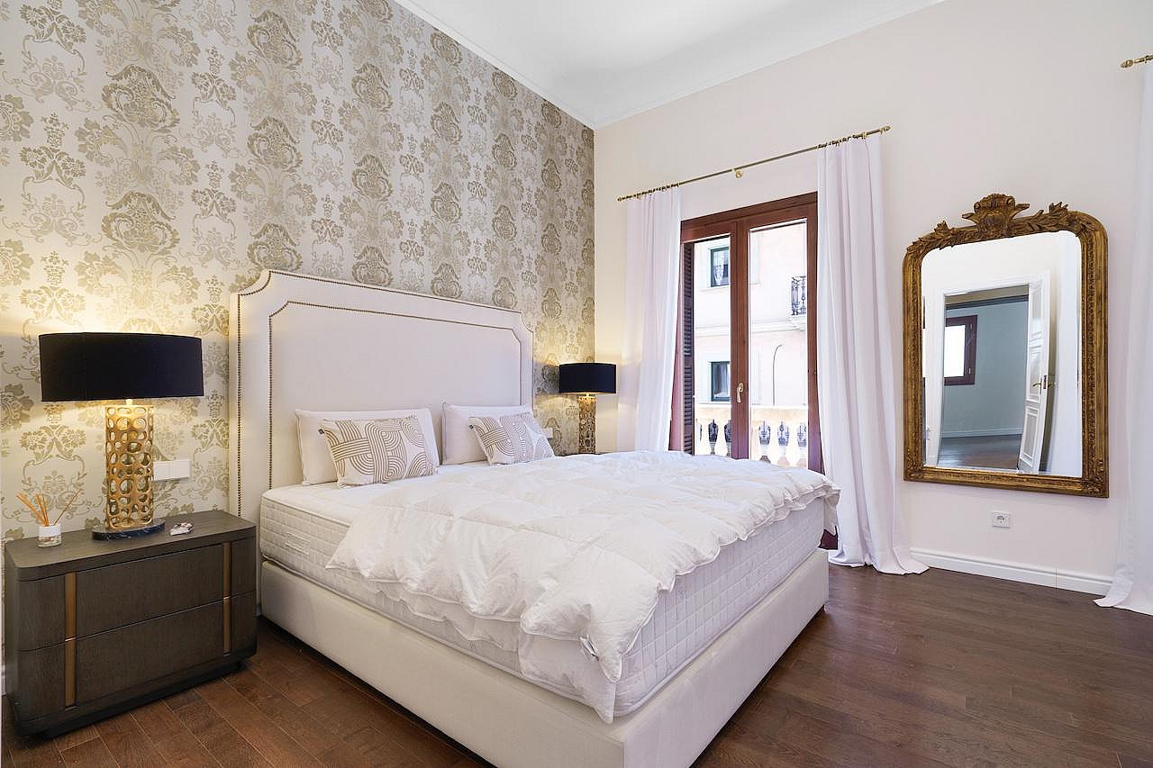 A master bedroom with a large bed against a damask patterned wall, dark wood floors, and a floor-length gold-framed mirror.