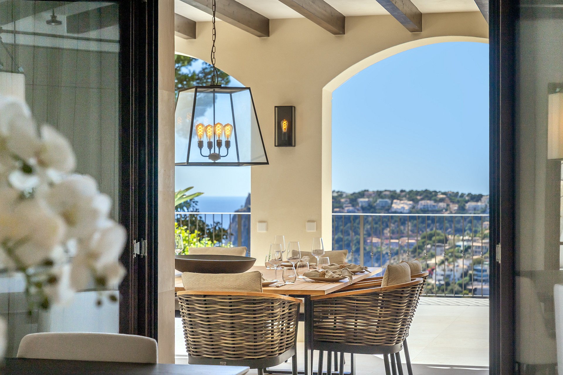 A dining table set with glassware on a covered terrace featuring wicker chairs, wood ceiling beams, and arched sea views.