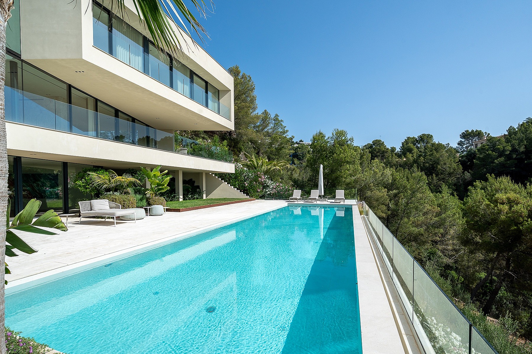 Wide exterior view of a white, multi-level modern villa featuring a long rectangular swimming pool, terrace, and trees.