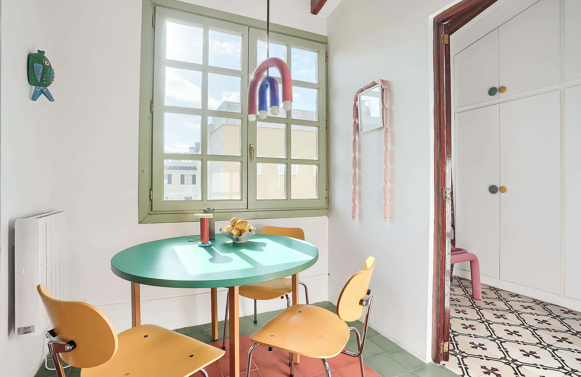 A small dining area with a round green table, wooden chairs, and a window overlooking city buildings in Santa Catalina.