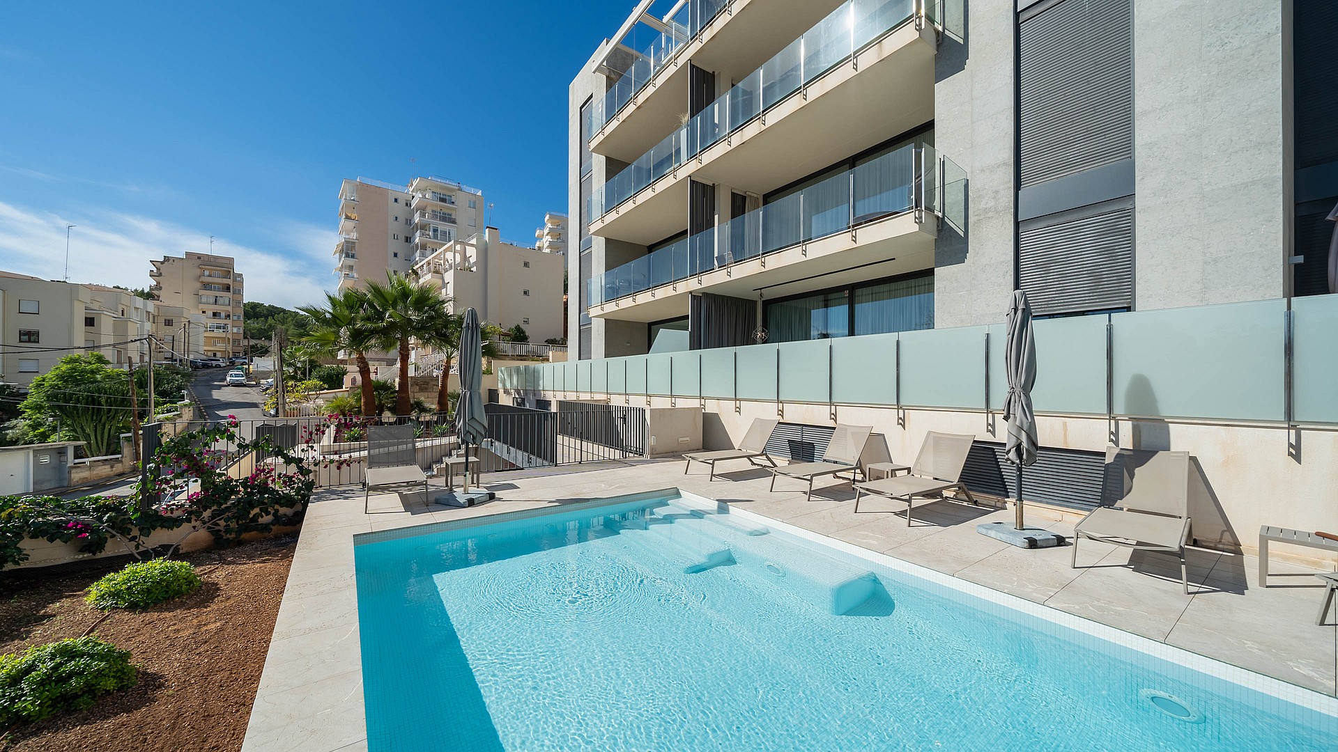A rectangular outdoor swimming pool with light blue water sits on a stone terrace in front of a modern multi-story apartment building with glass balcony railings.