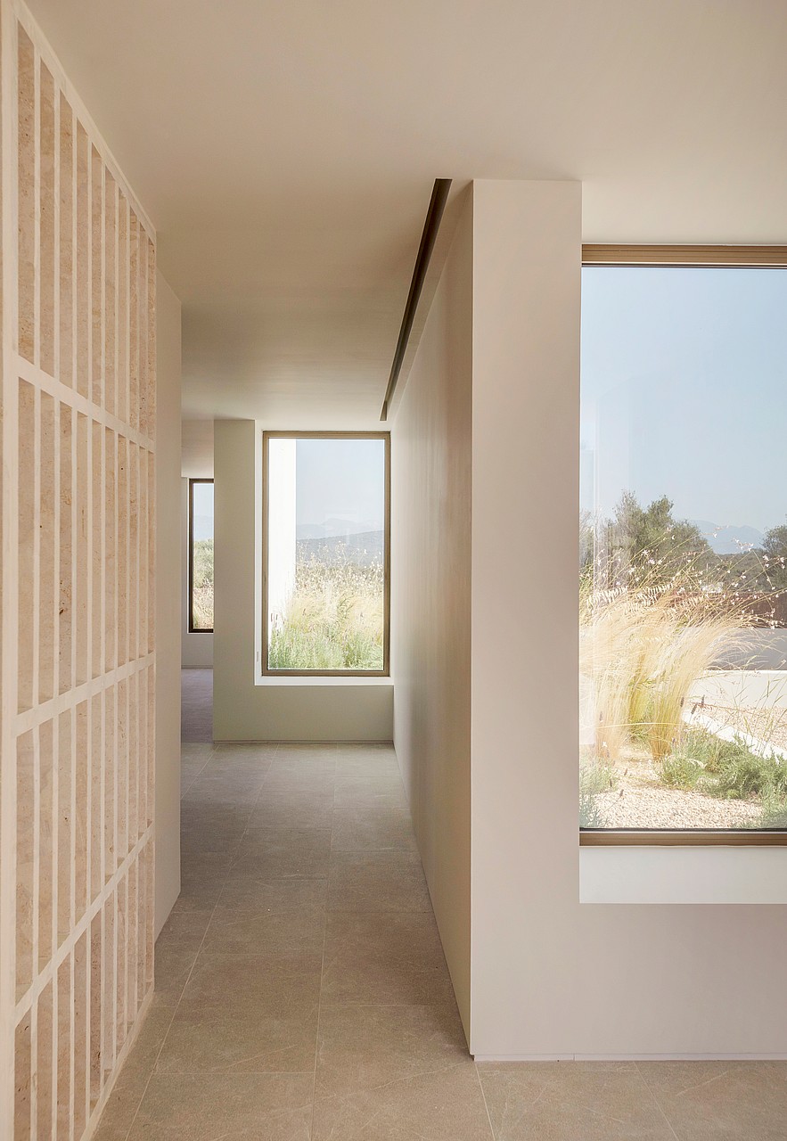 A sunlit hallway featuring light stone flooring, smooth plaster walls, and a vertical timber slatted screen beside large windows.