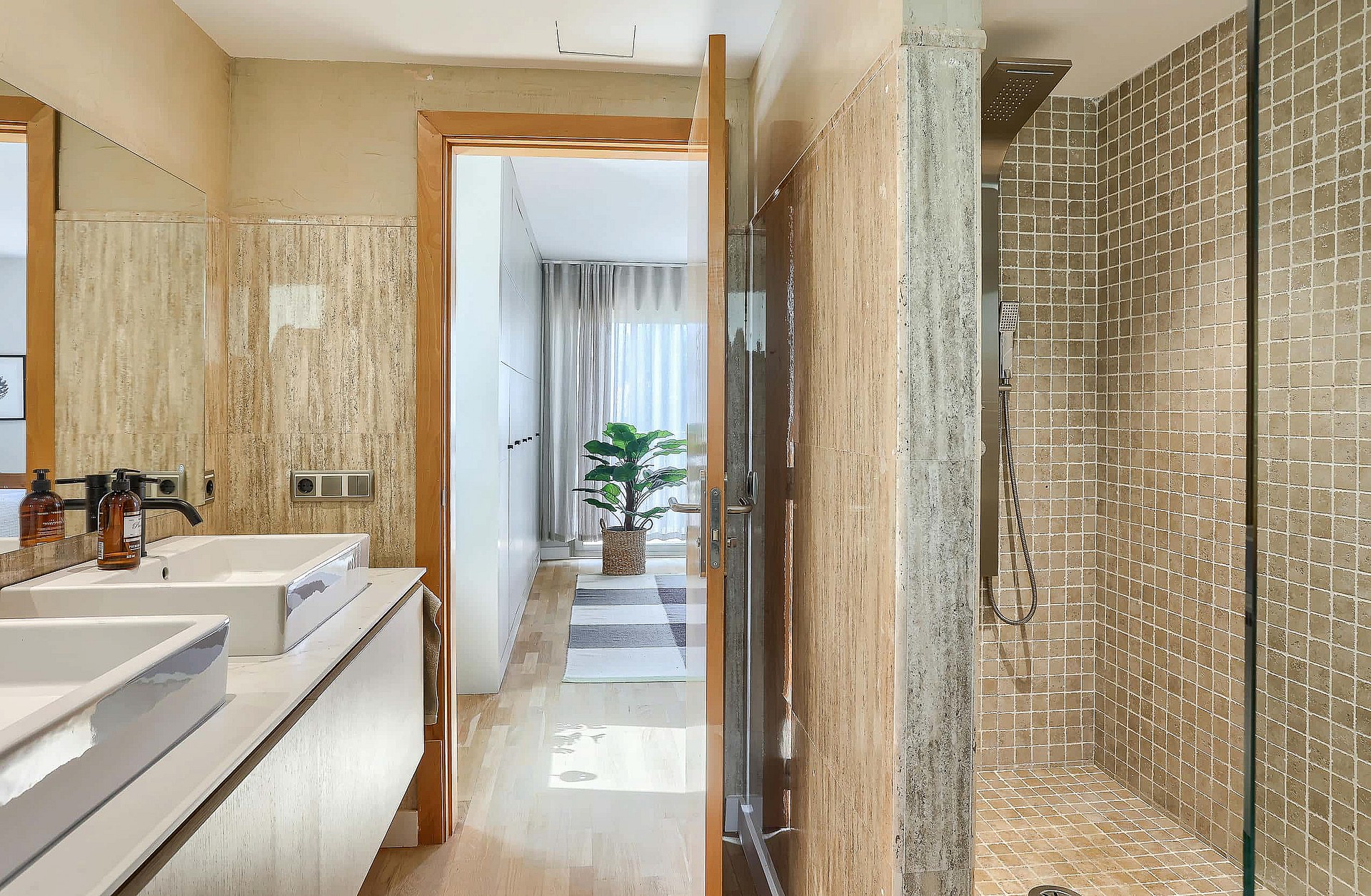 Modern bathroom featuring dual square sinks, stone-patterned tiled walls, and a glass-enclosed shower, leading into a bedroom.