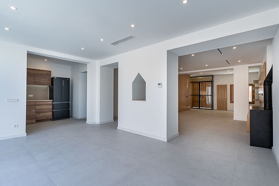 Wide shot of a contemporary interior with an open kitchen featuring dark wood cabinetry and a large refrigerator, transitioning to a bright, minimalist living space with grey tiled flooring and recessed ceiling lights.