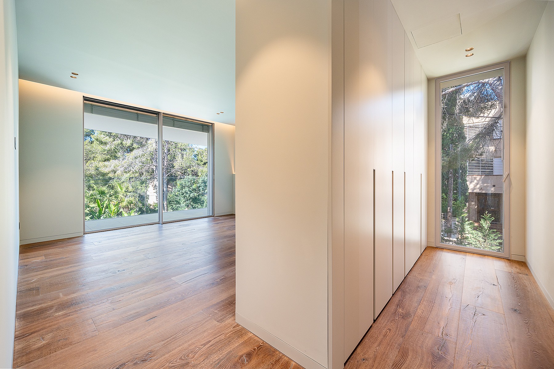 A minimalist dressing room corridor in a Santa Ponsa villa, featuring light oak wood floors, floor-to-ceiling white built-in wardrobes, and expansive glass windows overlooking greenery.