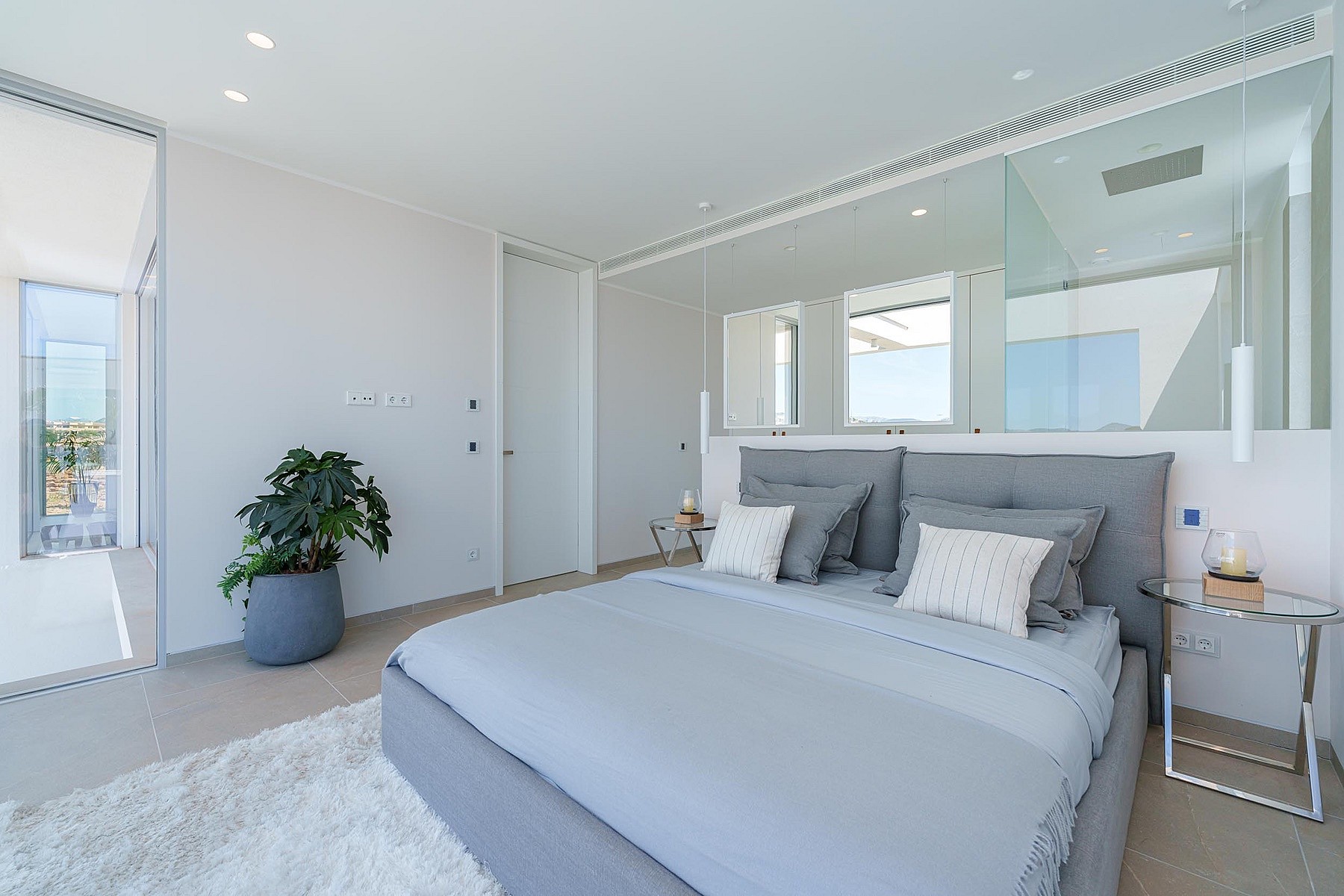 A modern bedroom with a grey upholstered bed, mirrored wall panels, and a shaggy white rug on a tiled floor.