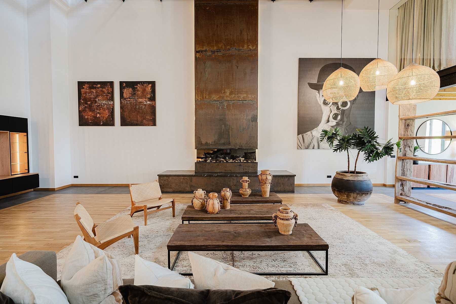 Spacious living room featuring a floor-to-ceiling oxidized metal fireplace, wooden coffee tables, low-profile seating, and woven pendant lights.