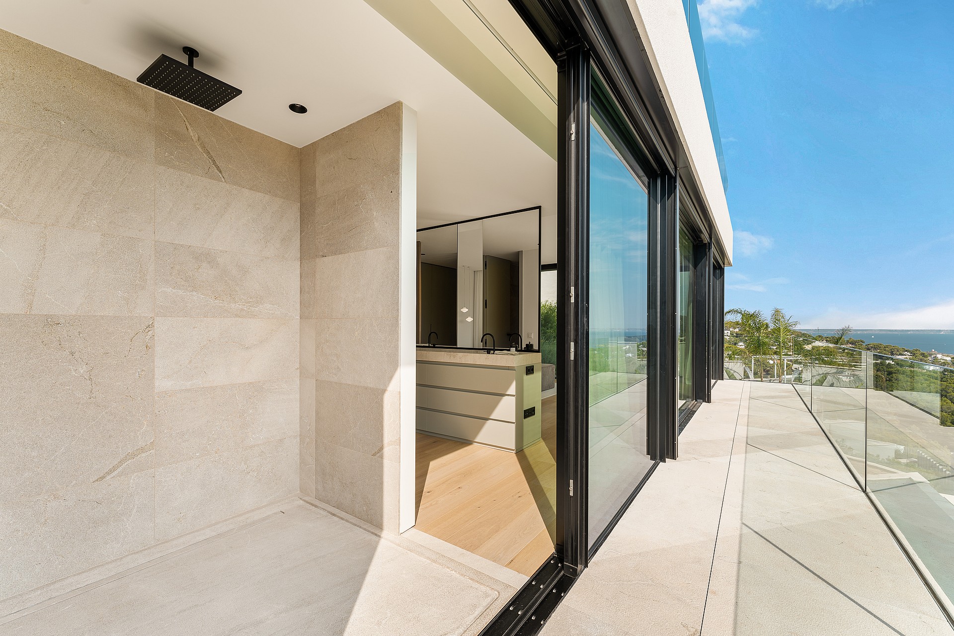 A wide shot looking from a stone-paved terrace into an interior room through sliding glass doors, with a clear blue sky above.
