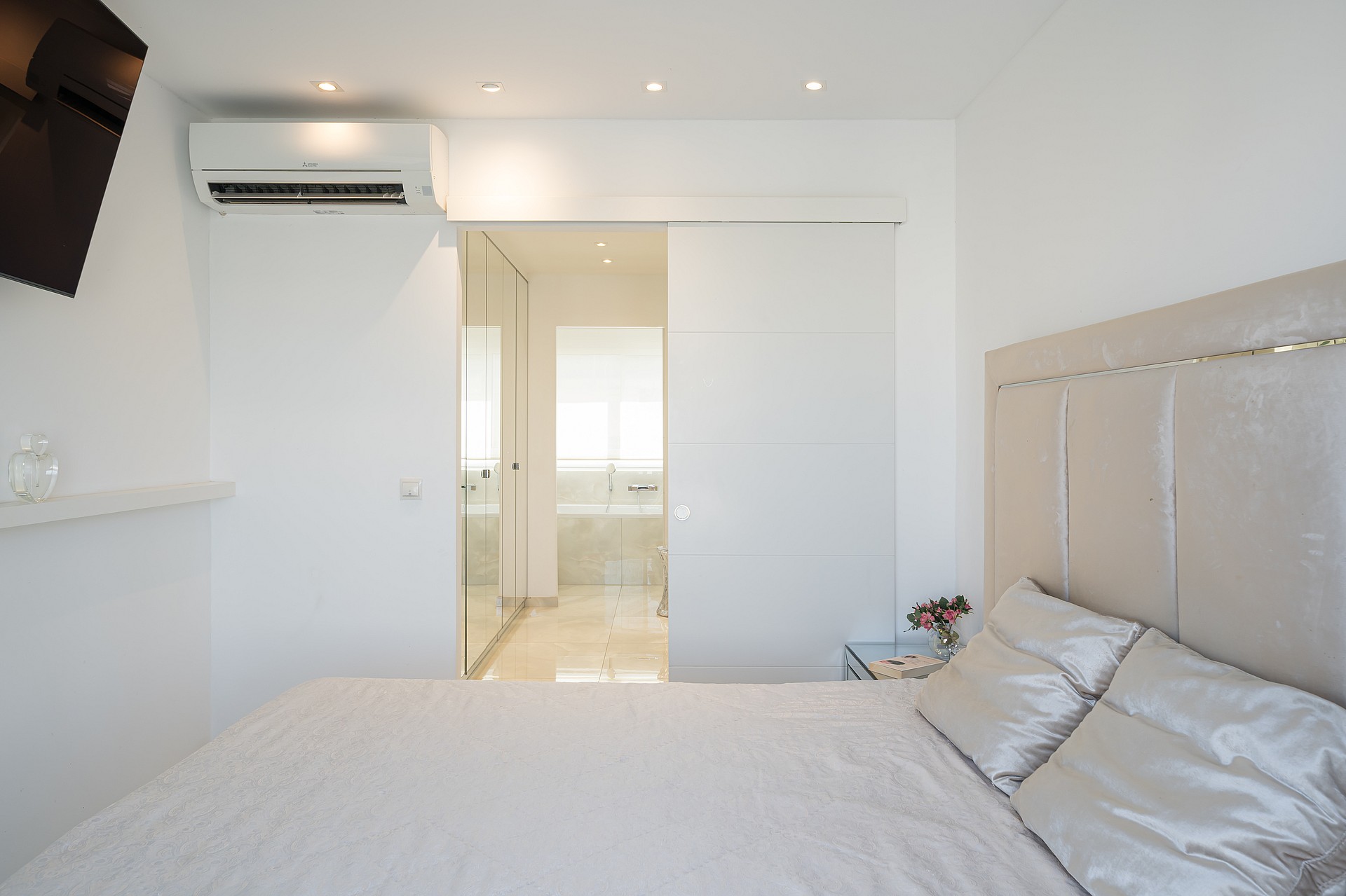 A modern bedroom with a neutral palette, a large bed with a padded headboard, and a sliding door leading to an en-suite bathroom.