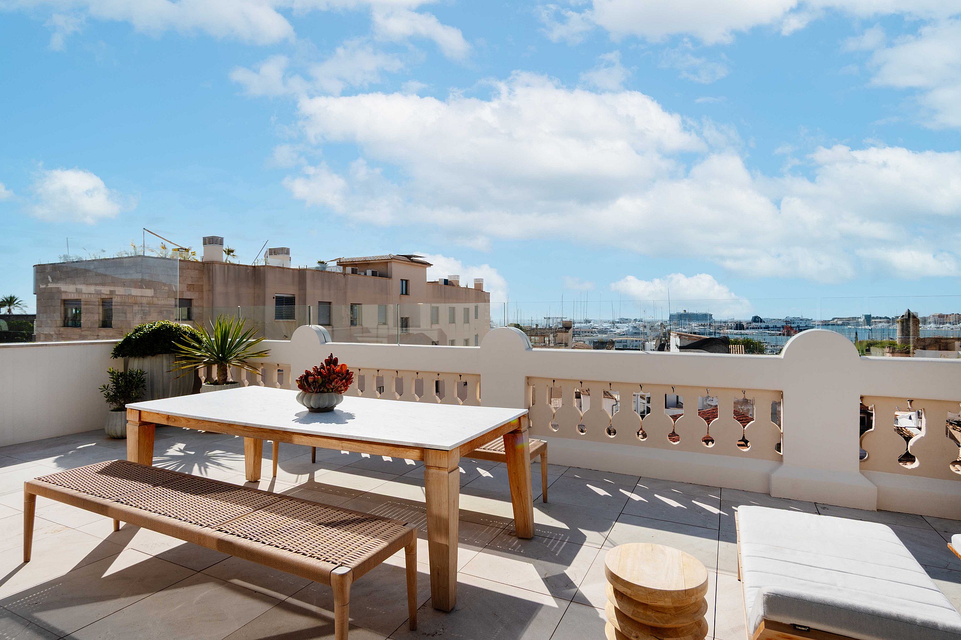 Rooftop terrace featuring a light wooden dining table with a bench, beige stone tiling, and traditional balustrade railings overlooking the Santa Catalina urban skyline.