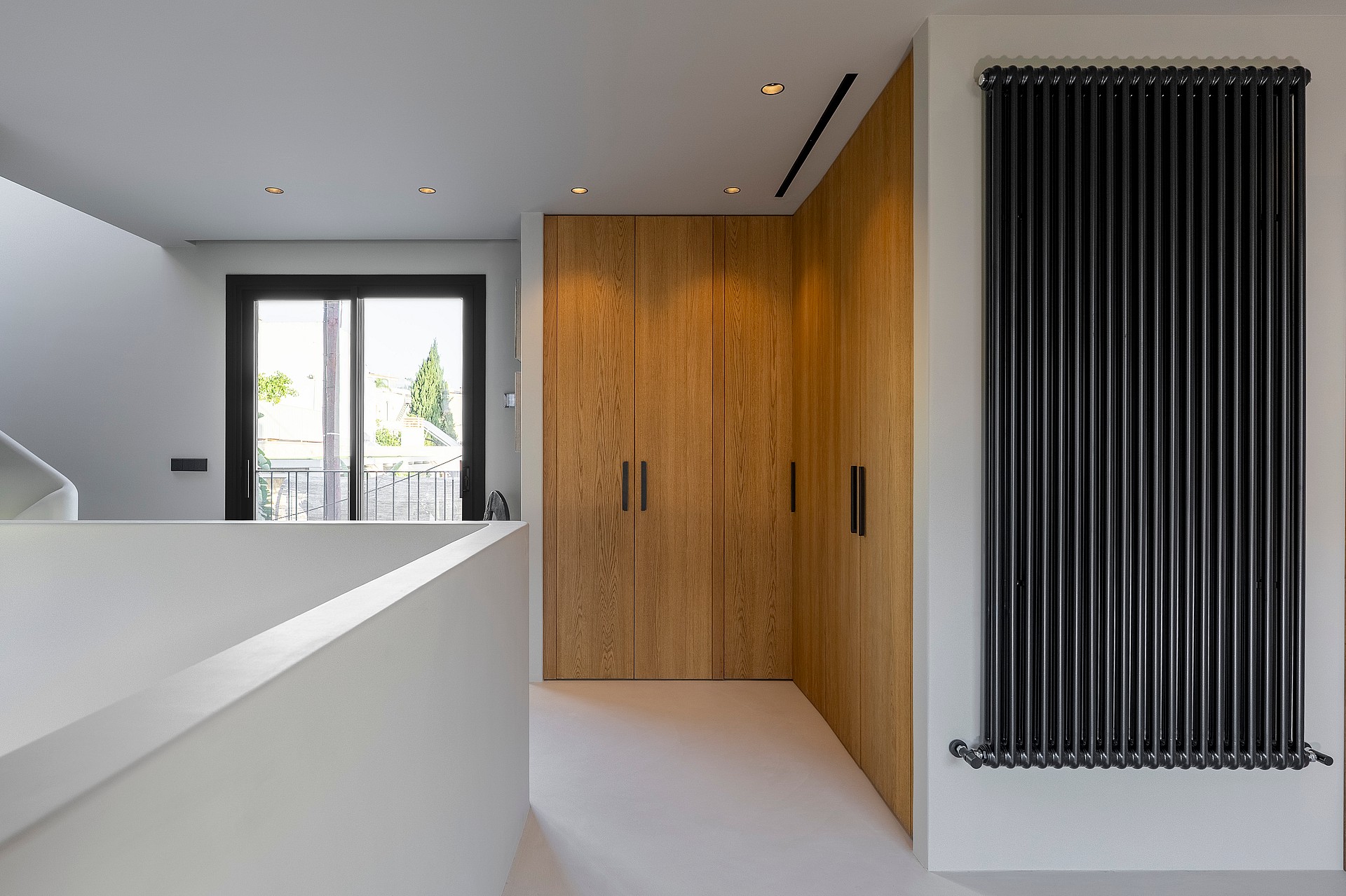 Modern hallway featuring custom oak wardrobes, a black vertical radiator, and natural light from a balcony door.