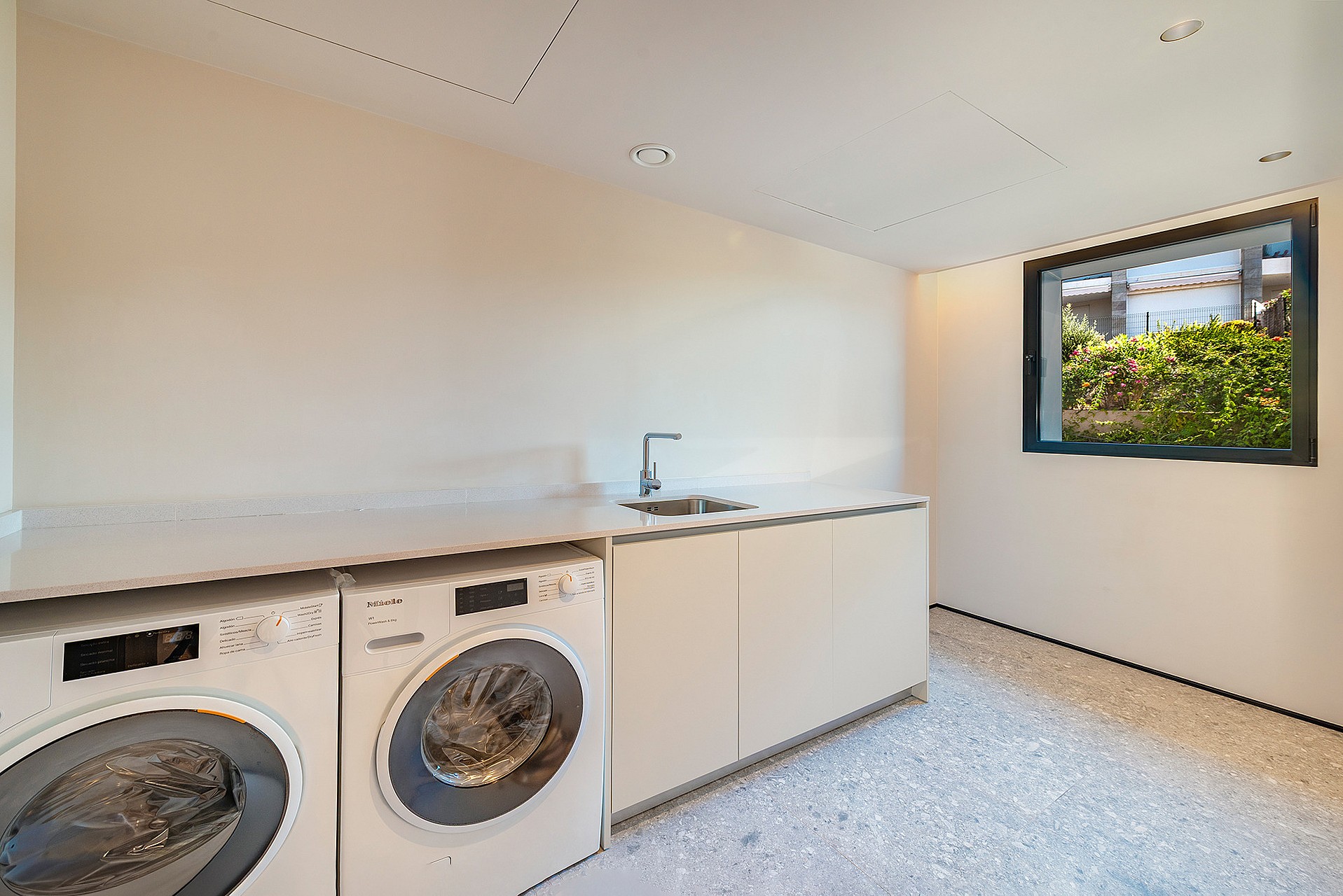 A minimalist laundry room featuring white front-loading appliances, a built-in sink, and speckled stone flooring.