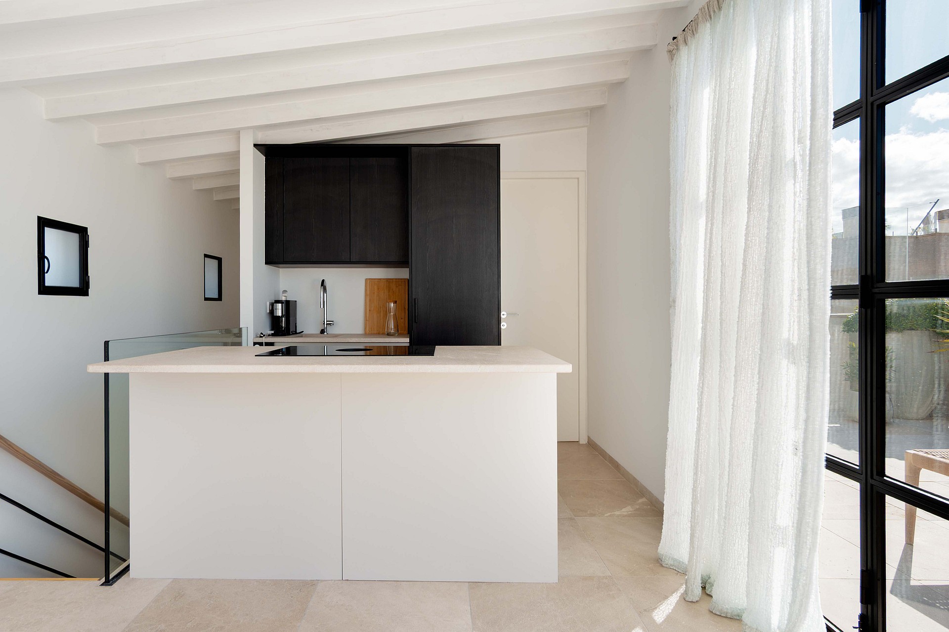 Modern kitchen with a white central island, dark cabinetry, and exposed white ceiling beams, adjacent to a staircase.