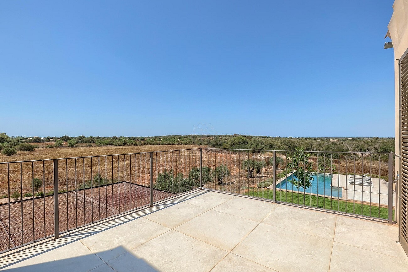 A stone-paved terrace with a metal railing overlooking a dry Mediterranean landscape, distant hills, and a swimming pool below.