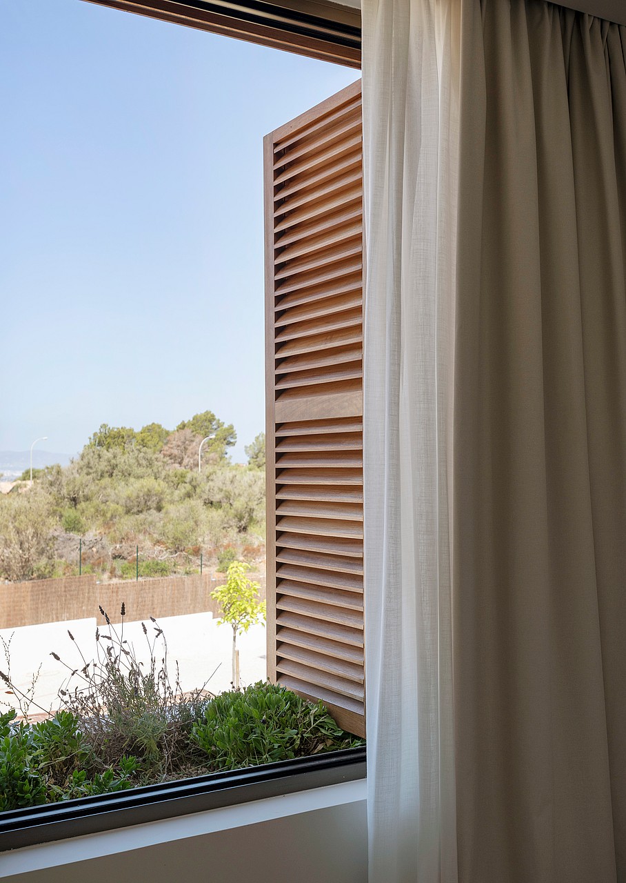 White curtains and wooden louvered shutters frame a window with an integrated planter box and green landscape beyond.
