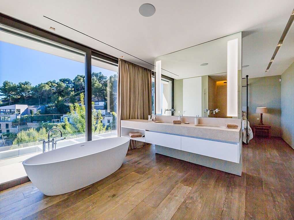 Modernt badrum med fristående badkar placerat vid glaspartier, dubbel kommod med integrerad belysning och trägolv.