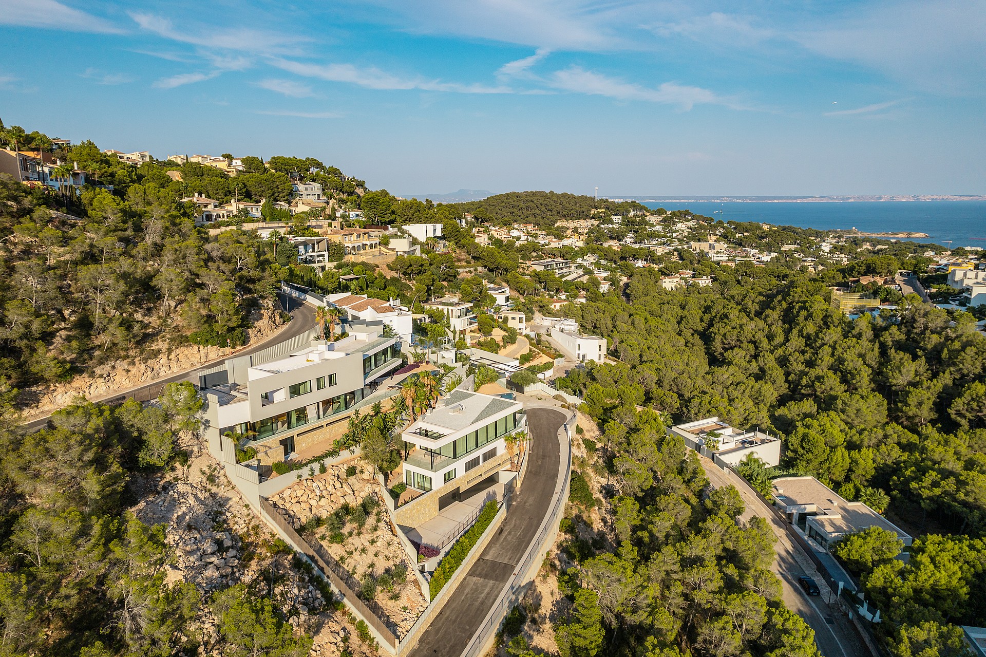 Aerial drone shot of a white contemporary hillside villa surrounded by pine trees, with a winding road and coastal horizon.