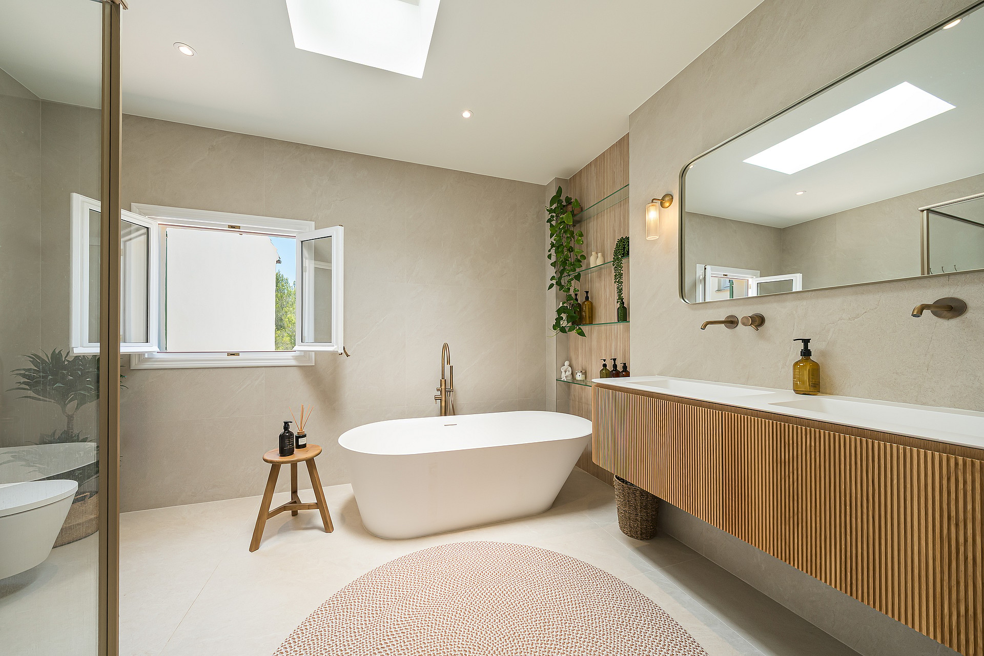 Contemporary bathroom with a freestanding oval bathtub, fluted wood vanity, brass fixtures, and natural light from a skylight.