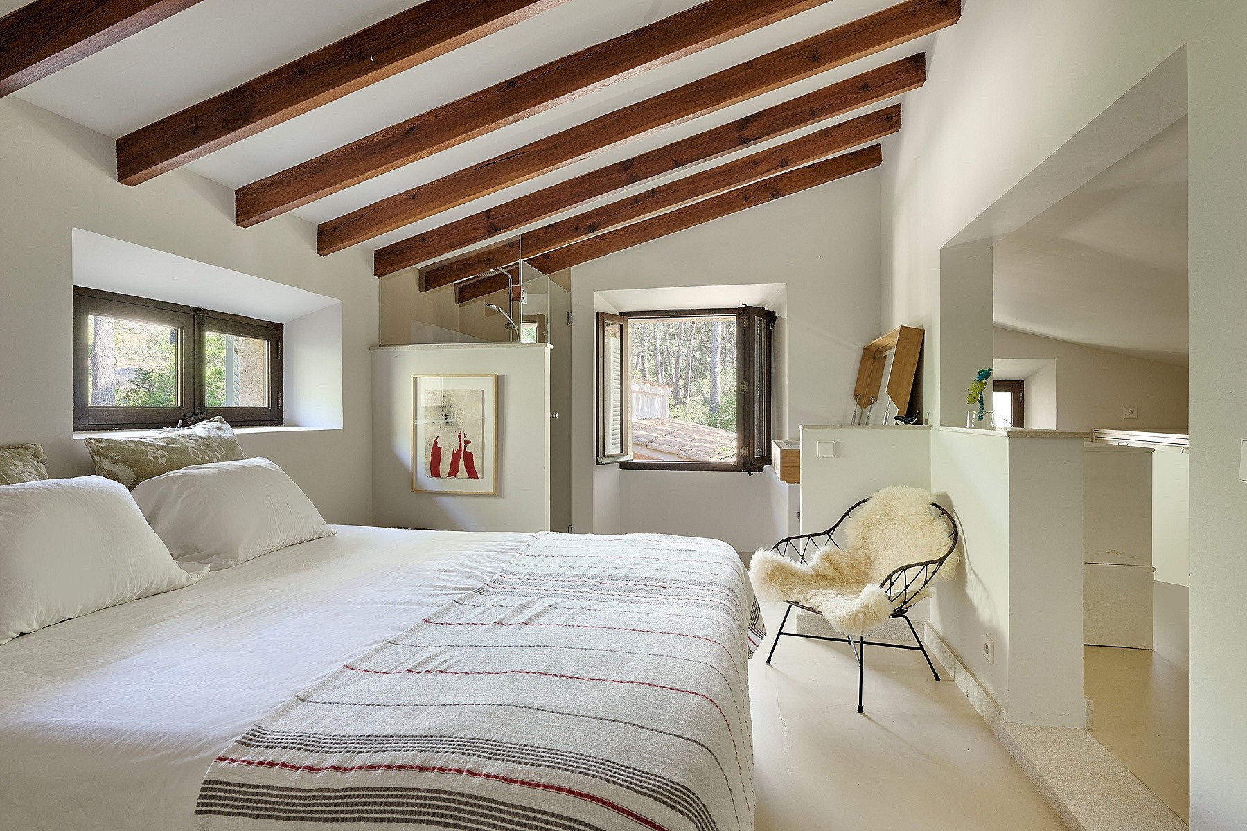Master bedroom with vaulted ceiling, exposed wooden beams, white bedding, and a decorative chair in an Alaro finca.