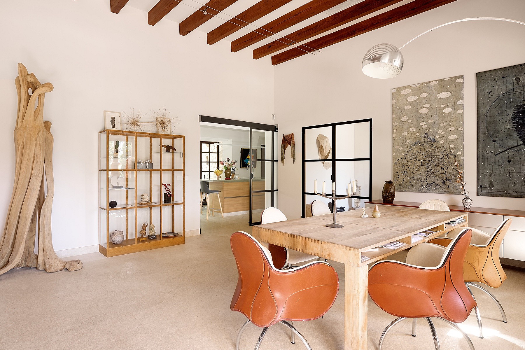 Dining area featuring a rustic wooden table, leather bucket chairs, and exposed timber ceiling beams in a spacious white room.