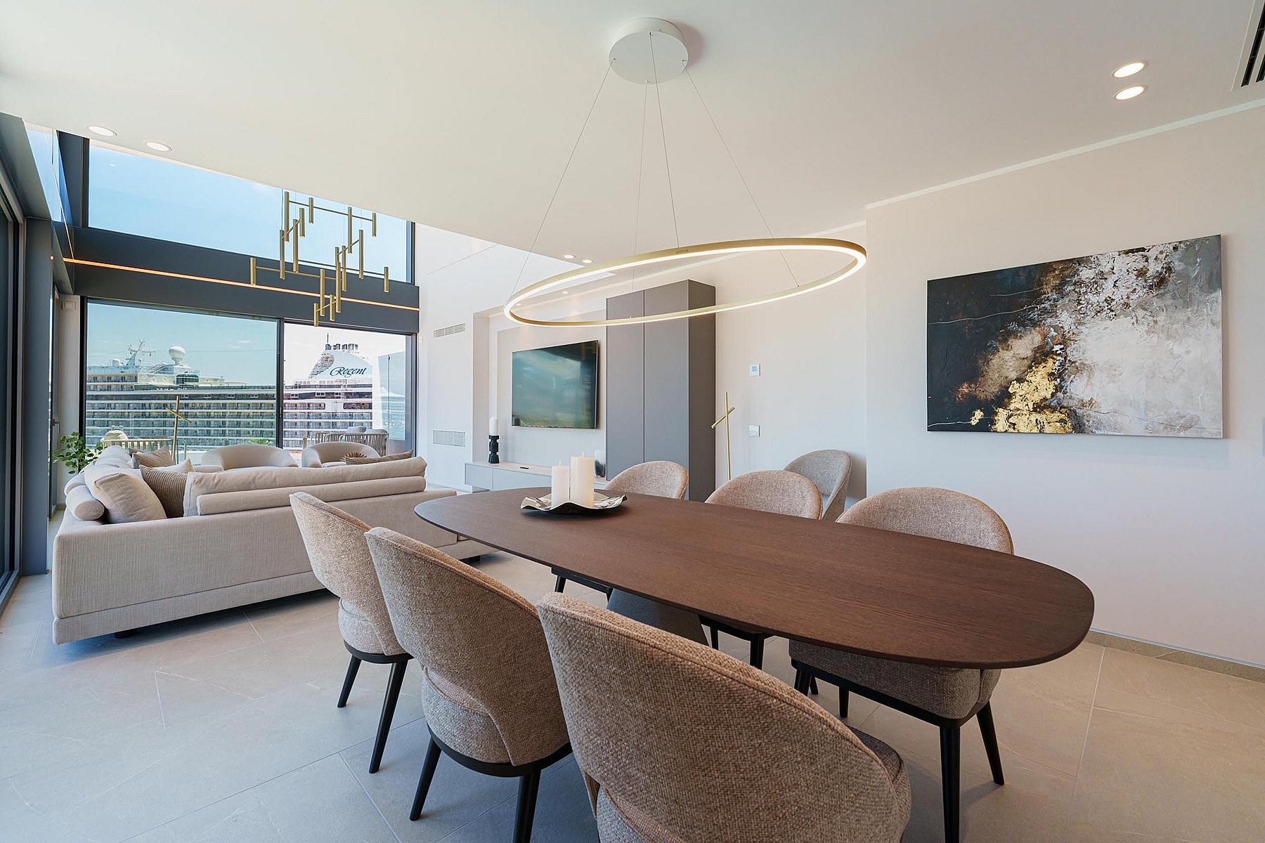 Comedor con mesa ovalada de madera, sillas tapizadas y lámpara circular moderna, abierto hacia el mar.