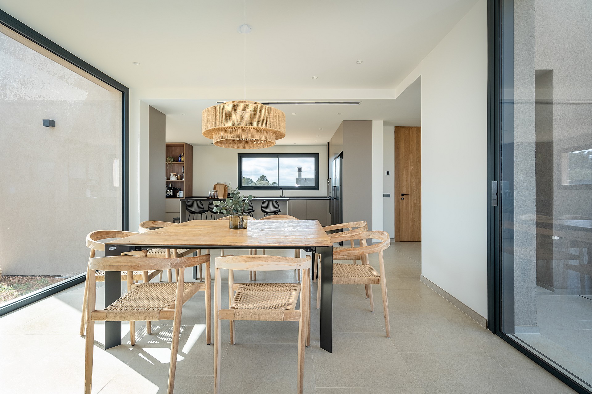 Dining room with a wooden table, woven chairs, and a large pendant light, framed by glass sliding doors and stone flooring.
