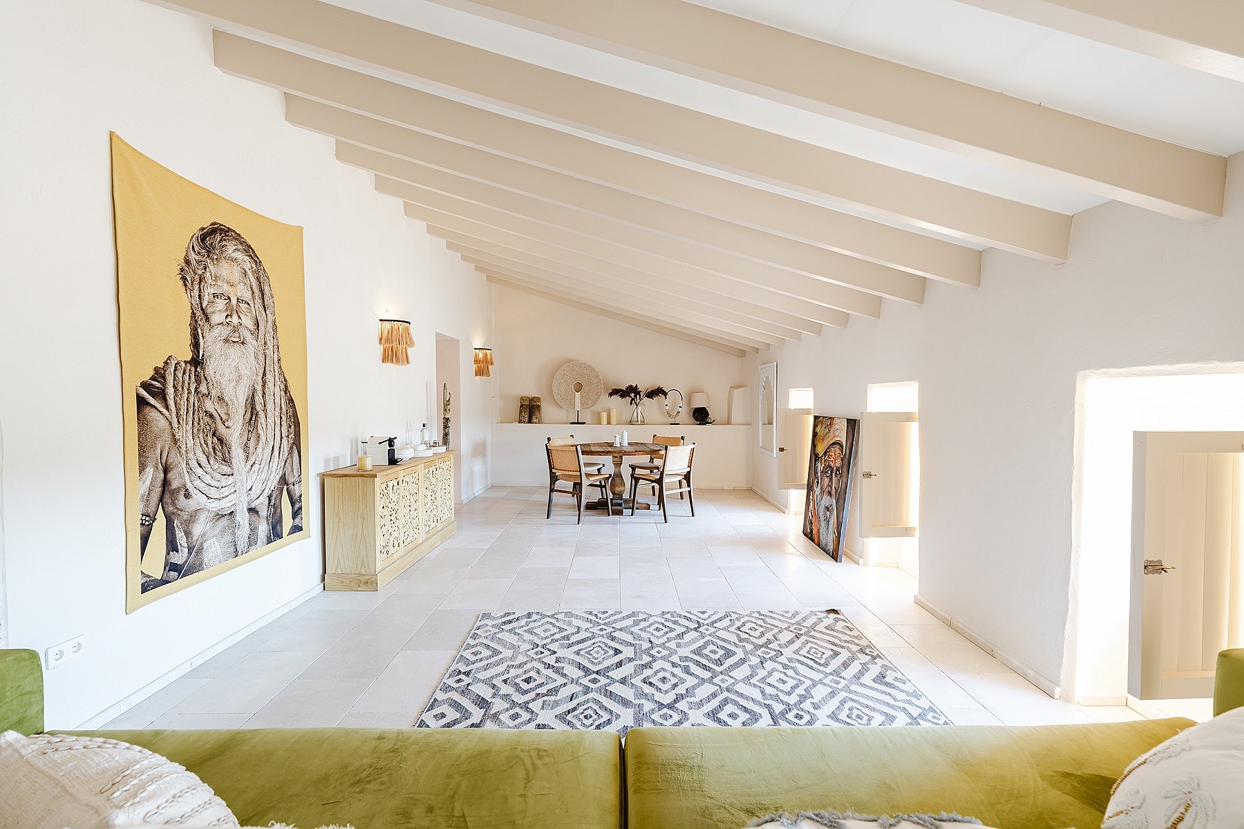 A spacious living room in a Sencelles townhouse featuring high vaulted ceilings with exposed cream beams, white walls, and a large artistic wall hanging.