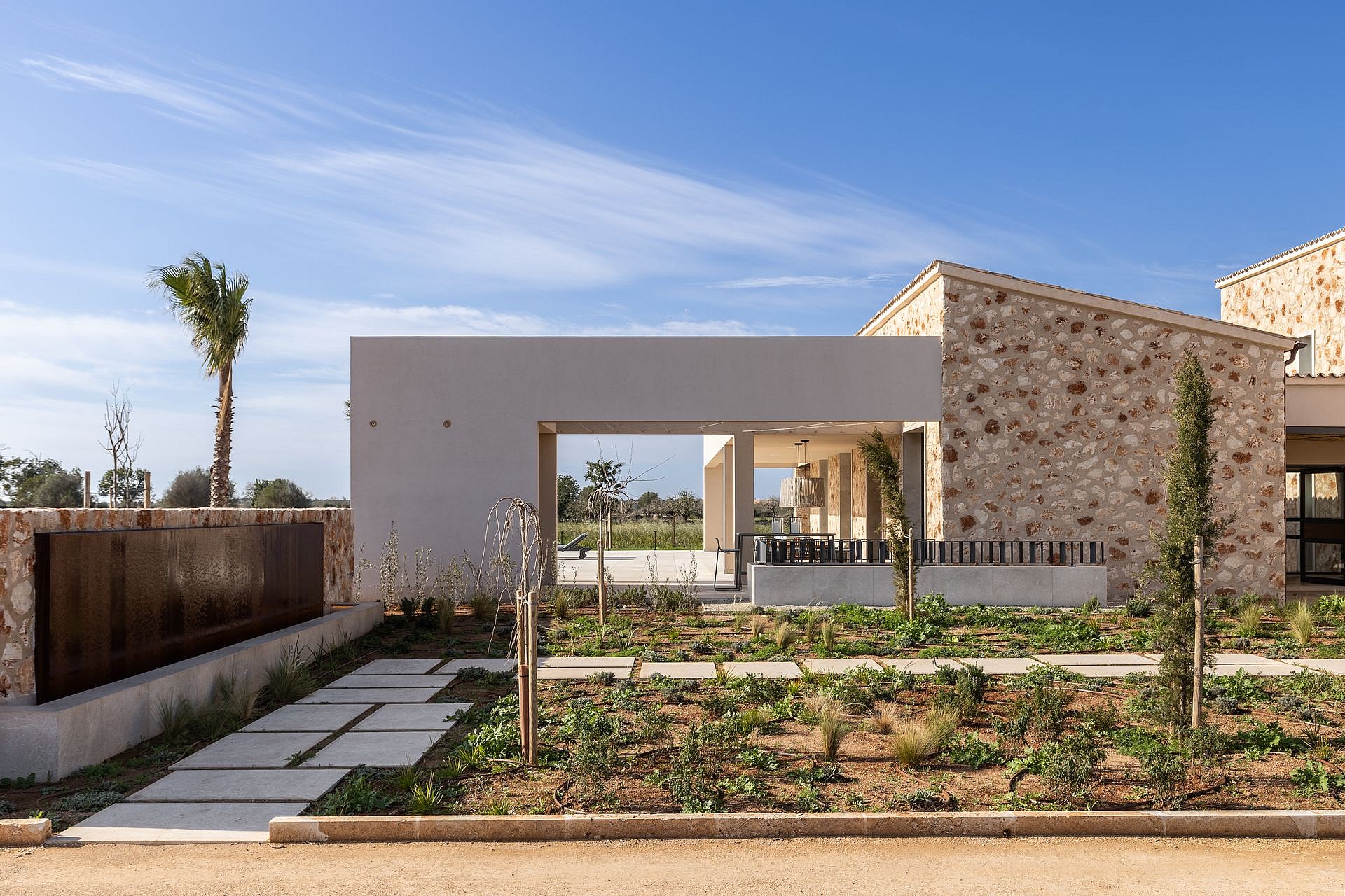 Exterior view of a modern finca featuring traditional natural stone walls, a flat concrete overhang, and a landscaped garden.