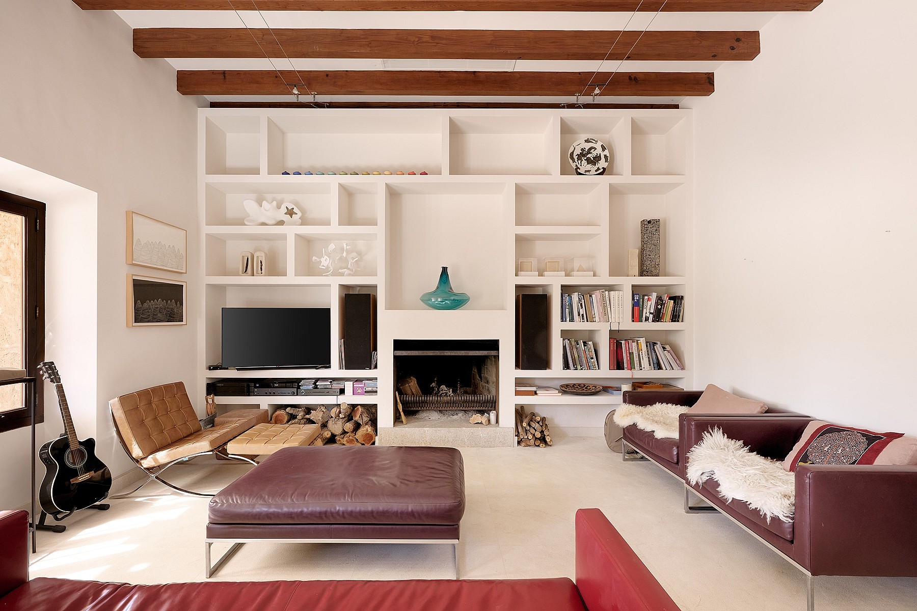 A living area with white walls, wooden ceiling beams, and a floor-to-ceiling white shelving unit built around a fireplace.