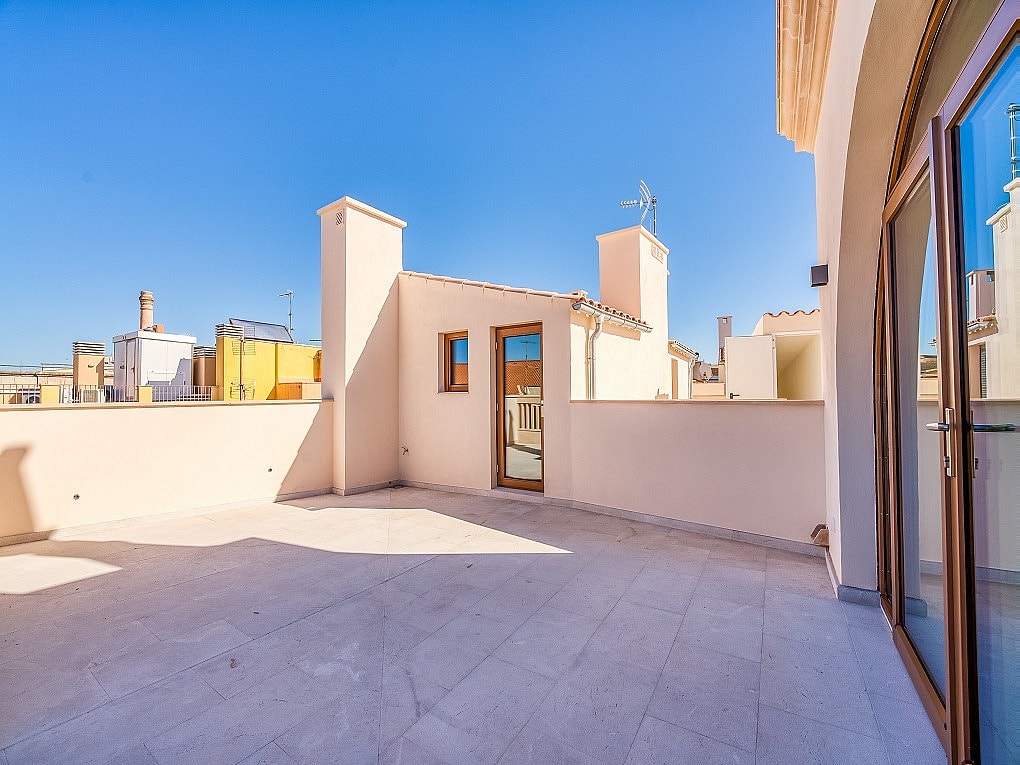 An open stone-paved rooftop terrace with light-colored walls and glass doors under a clear blue sky, overlooking city buildings.