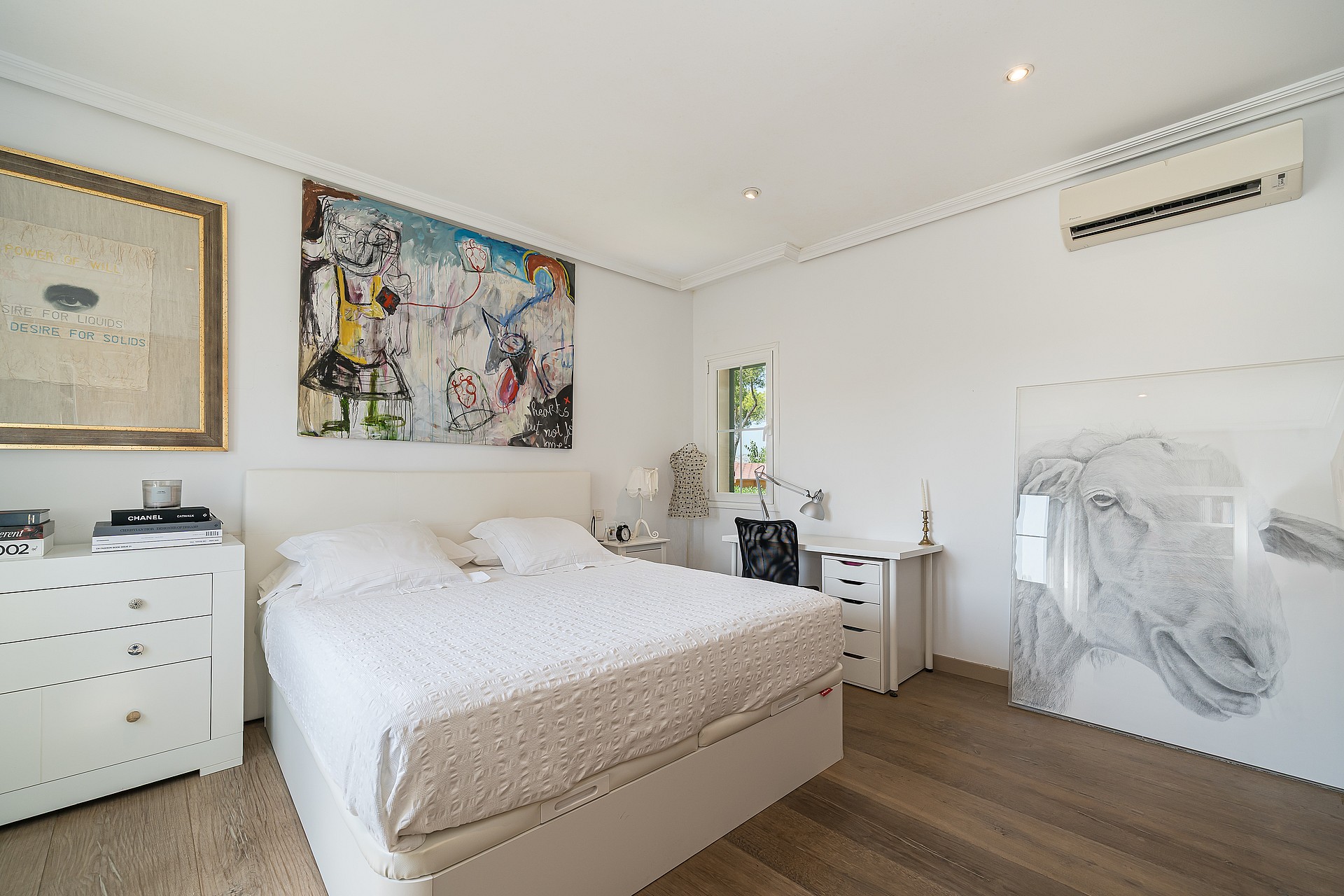 A bright bedroom with white walls, a king-size bed, a large abstract painting, and light-toned hardwood flooring.