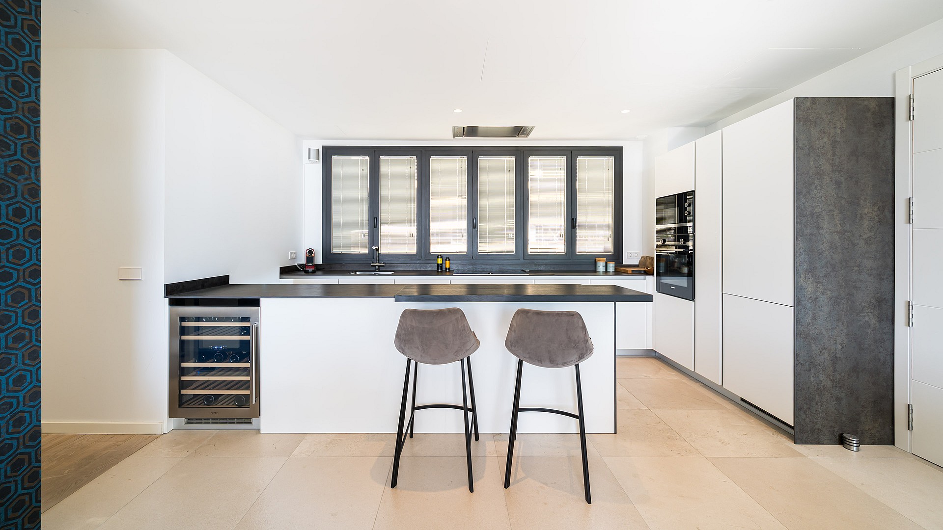 Modern kitchen featuring a white island with two stools, dark countertops, and a built-in wine fridge against a bright window.
