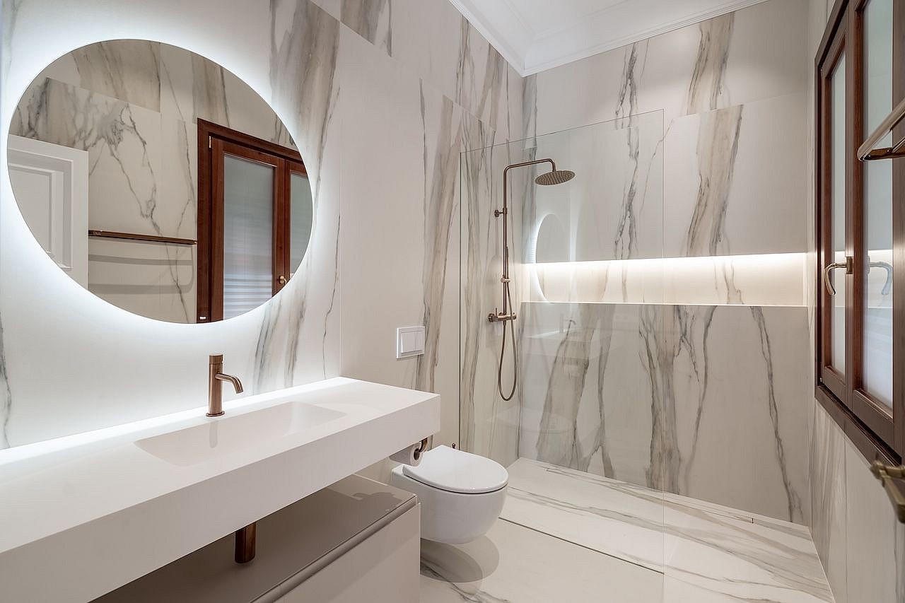 A contemporary bathroom featuring floor-to-ceiling white marble with grey veining, a floating vanity, and a circular LED-lit mirror.
