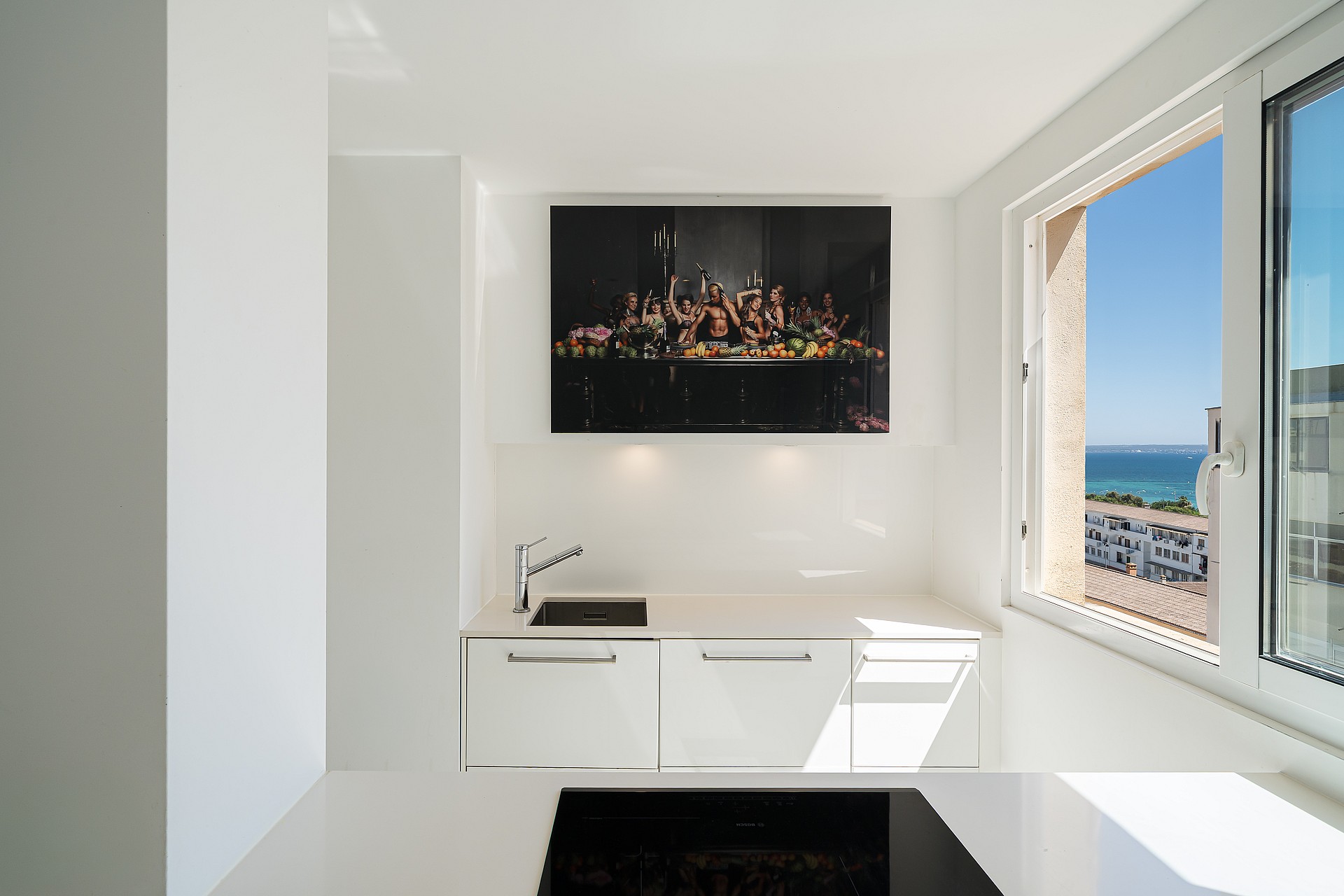 Modern white kitchen featuring a countertop, sink, and high-contrast artwork, positioned next to a large window overlooking the city.