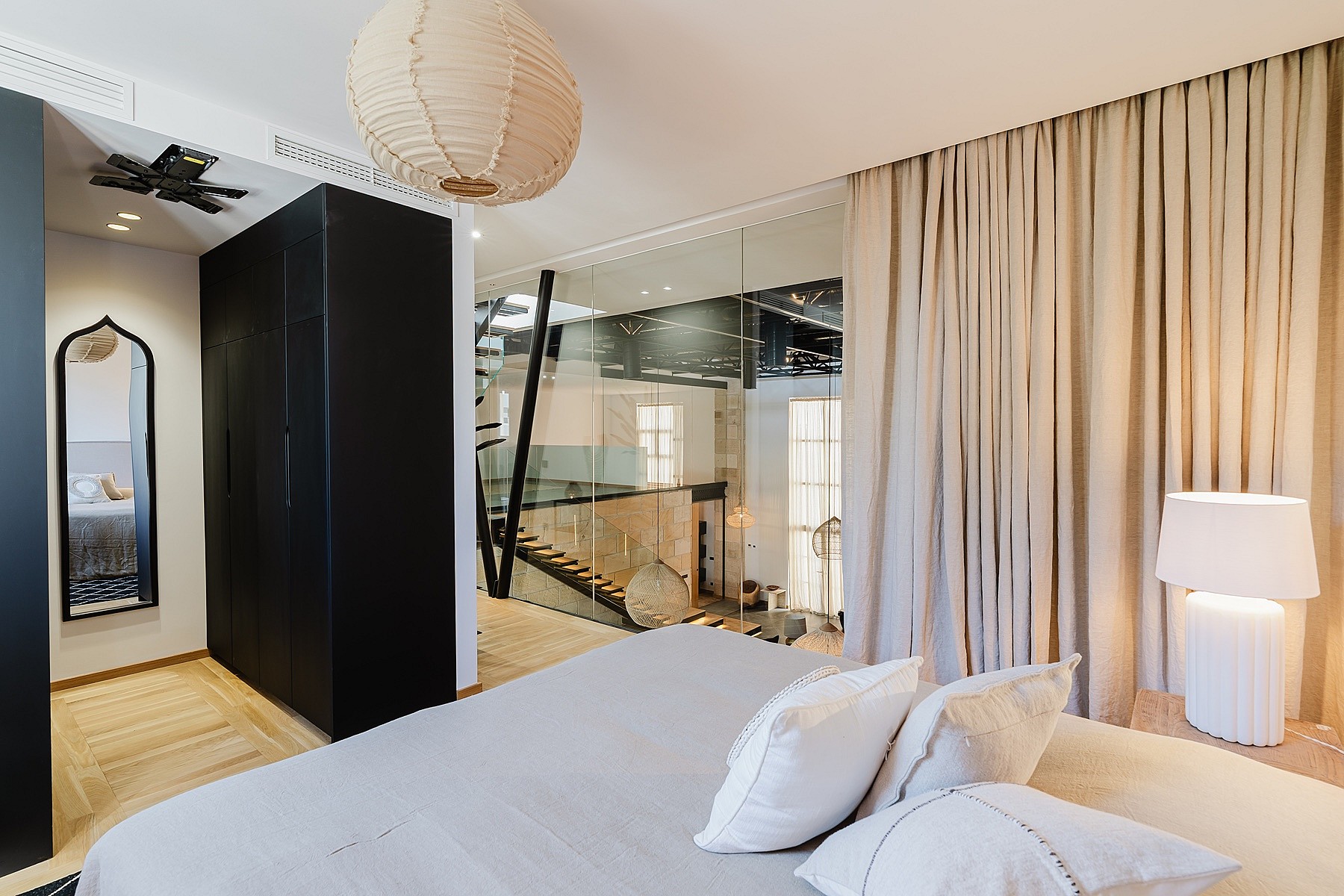 Modern master bedroom featuring a large bed, dark matte wardrobe, and glass walls separating the space from an internal staircase.