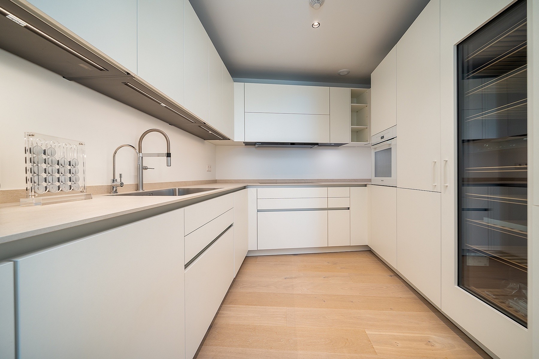 A sleek galley-style kitchen with white handleless cabinetry, light oak wood flooring, and a glass-fronted wine refrigerator.