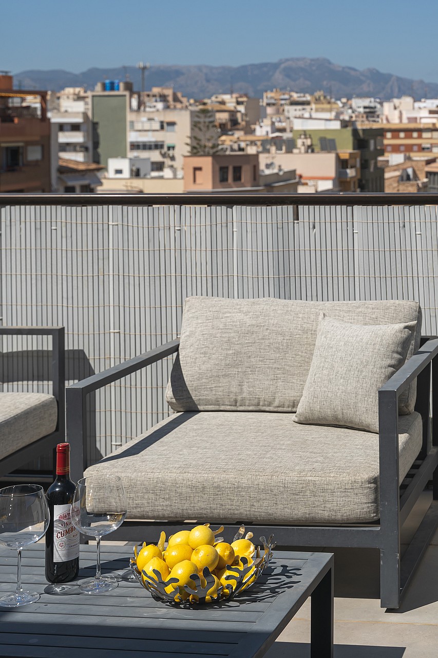 A modern outdoor lounge chair with grey fabric cushions sits on a terrace with a metal privacy screen overlooking a city.