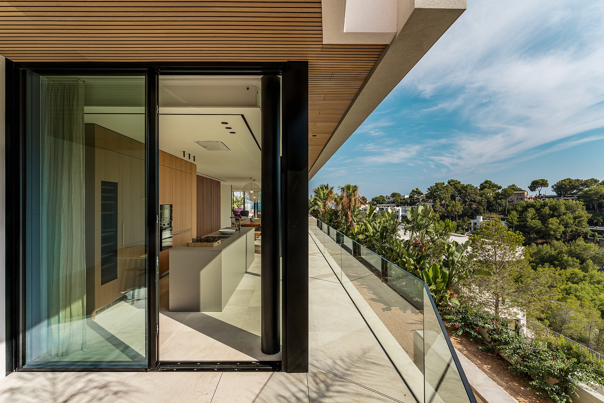 Wide angle view from a modern kitchen looking out onto a long stone terrace with a frameless glass balustrade and greenery.