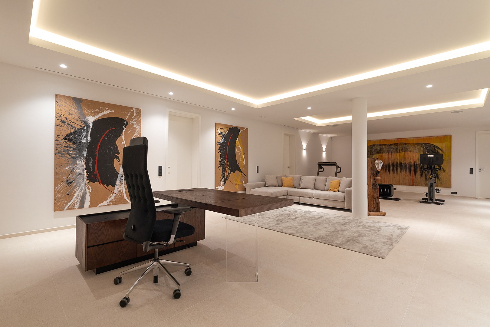 Interior view of an open-plan office space featuring a dark wood desk, contemporary paintings on white walls, and a recessed lounge corner with a grey sofa and gym equipment.
