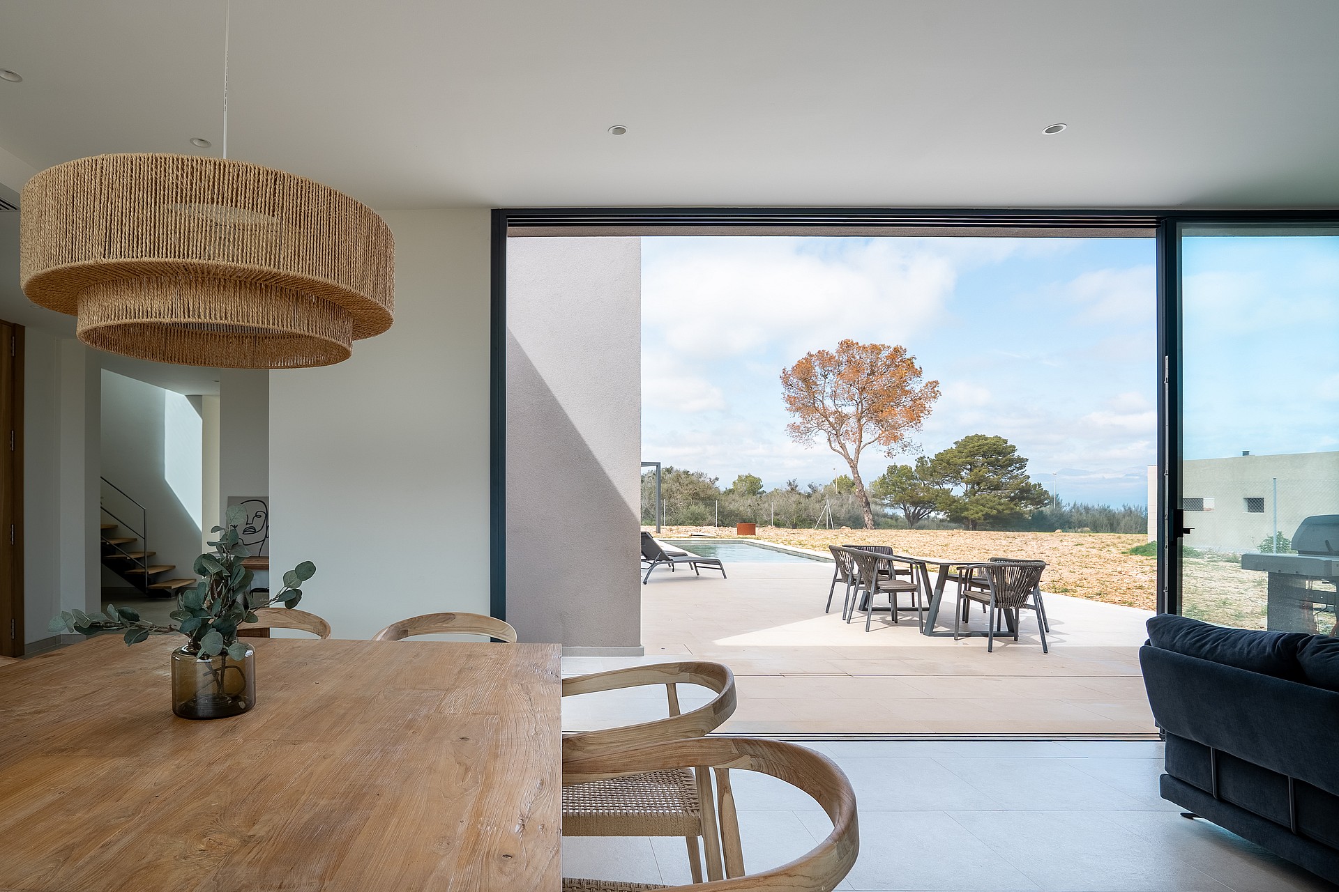 Dining area with a large wooden table and textured pendant light opening onto a terrace with a stone wall and mountain views.