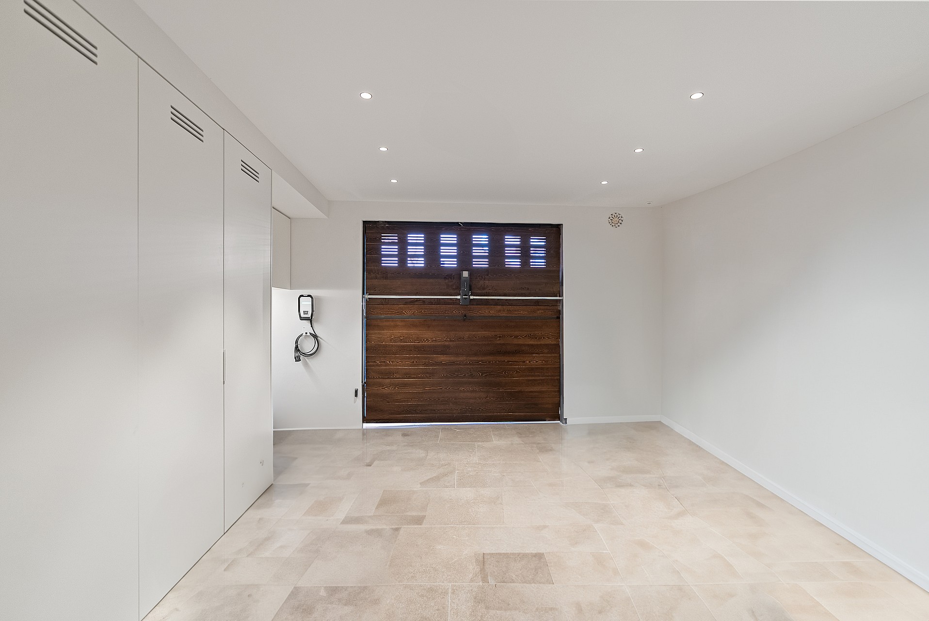 Modern garage featuring polished stone floors, white minimalist cabinets, a dark wood door, and a wall-mounted EV charger.