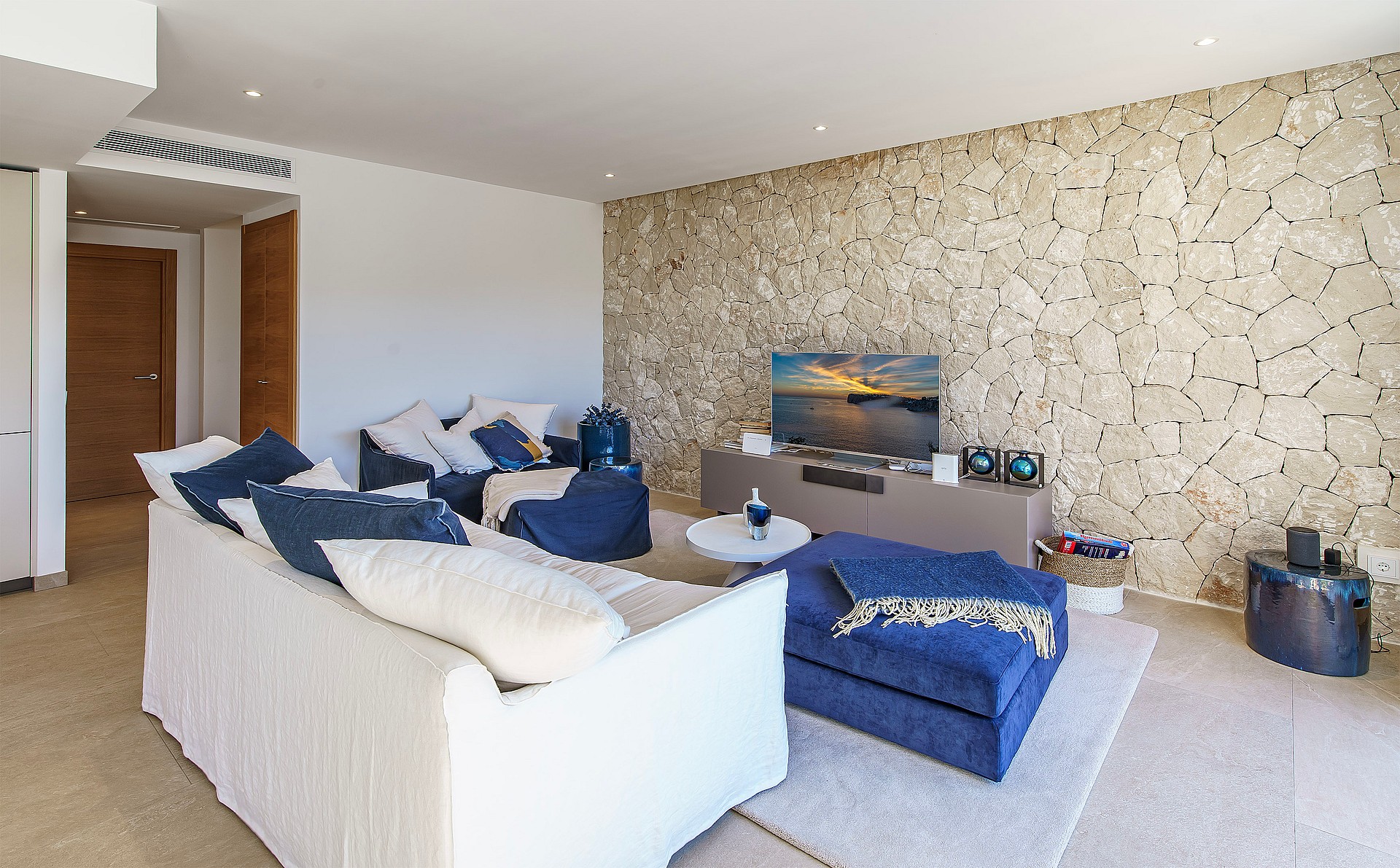 Modern living room featuring a textured natural stone wall, white sofa, blue armchairs, and neutral tiled flooring.