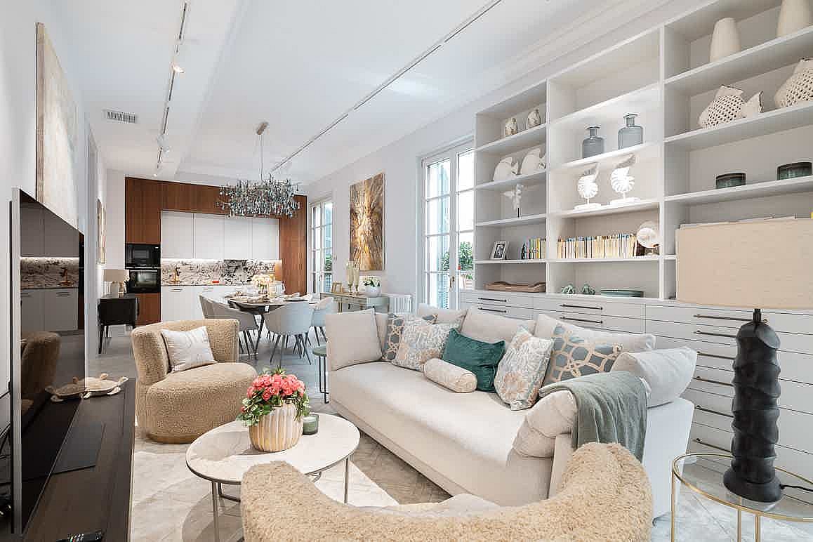 Open-plan living room with white custom built-in shelving, a neutral beige sofa, and two plush armchairs facing a kitchen area.