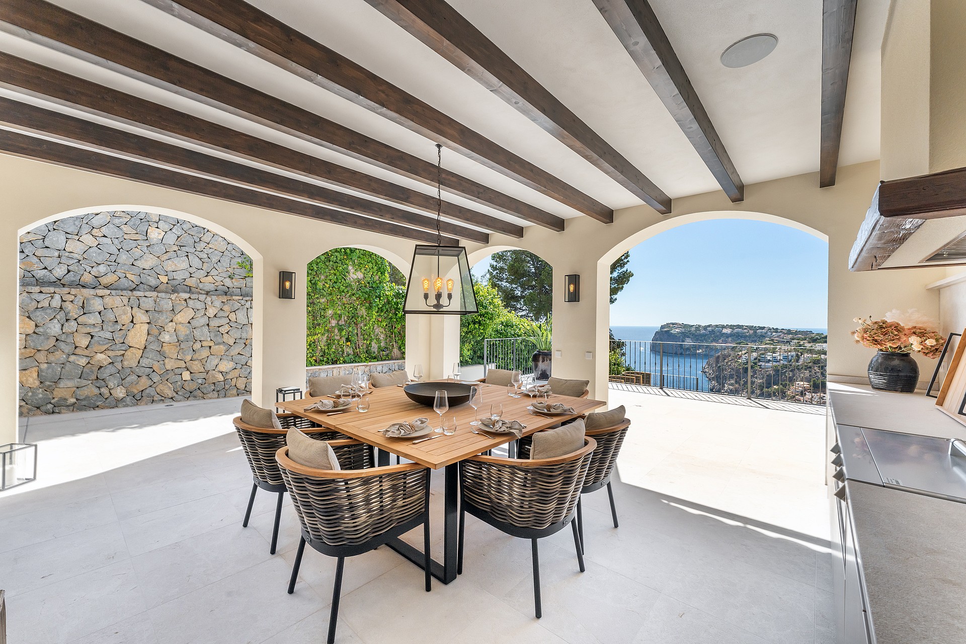 Covered terrace with dark wooden ceiling beams, a large square table, woven chairs, and views of a sea bay through arches.