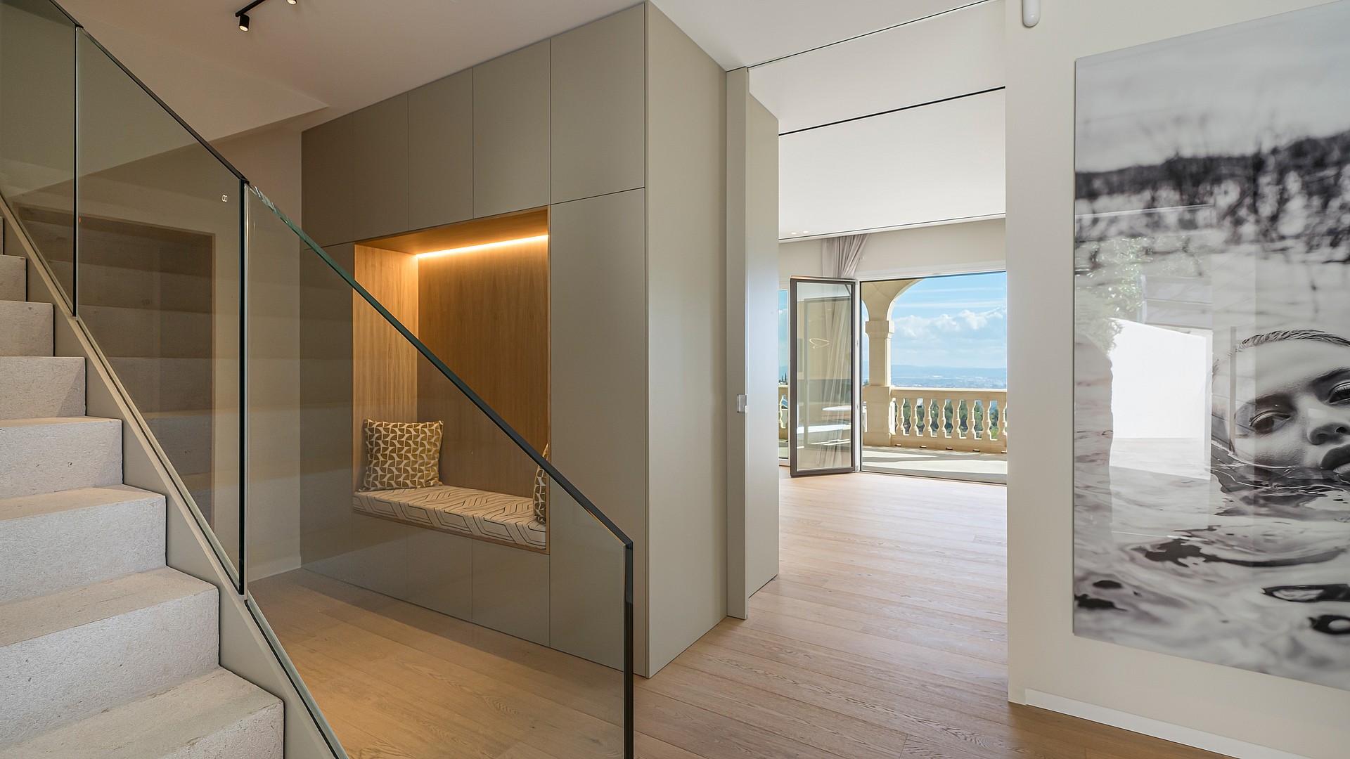 A modern staircase with a frameless glass railing leads past a built-in wall niche with a padded bench and warm lighting.