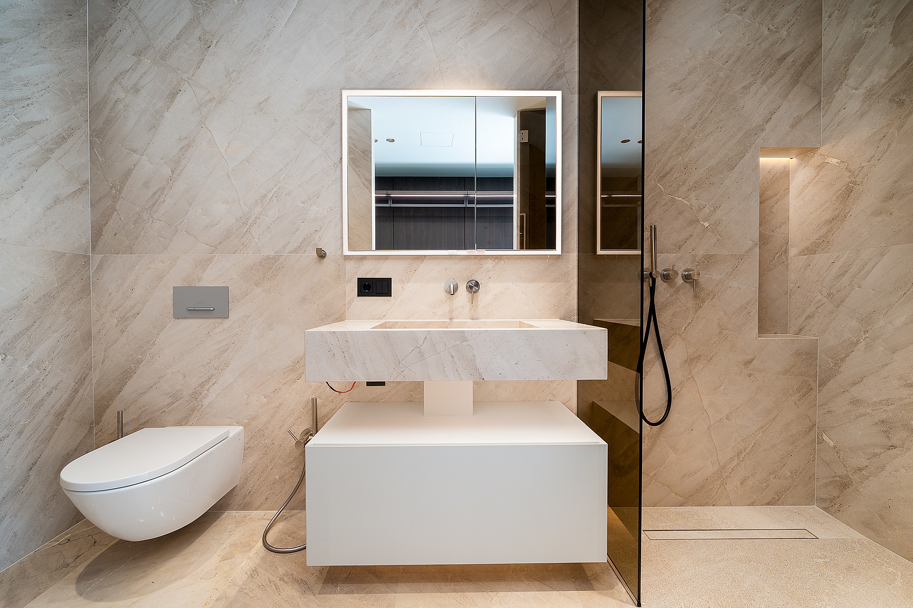 A modern bathroom featuring beige veined marble walls, a wall-hung white vanity, and a glass-enclosed walk-in shower.