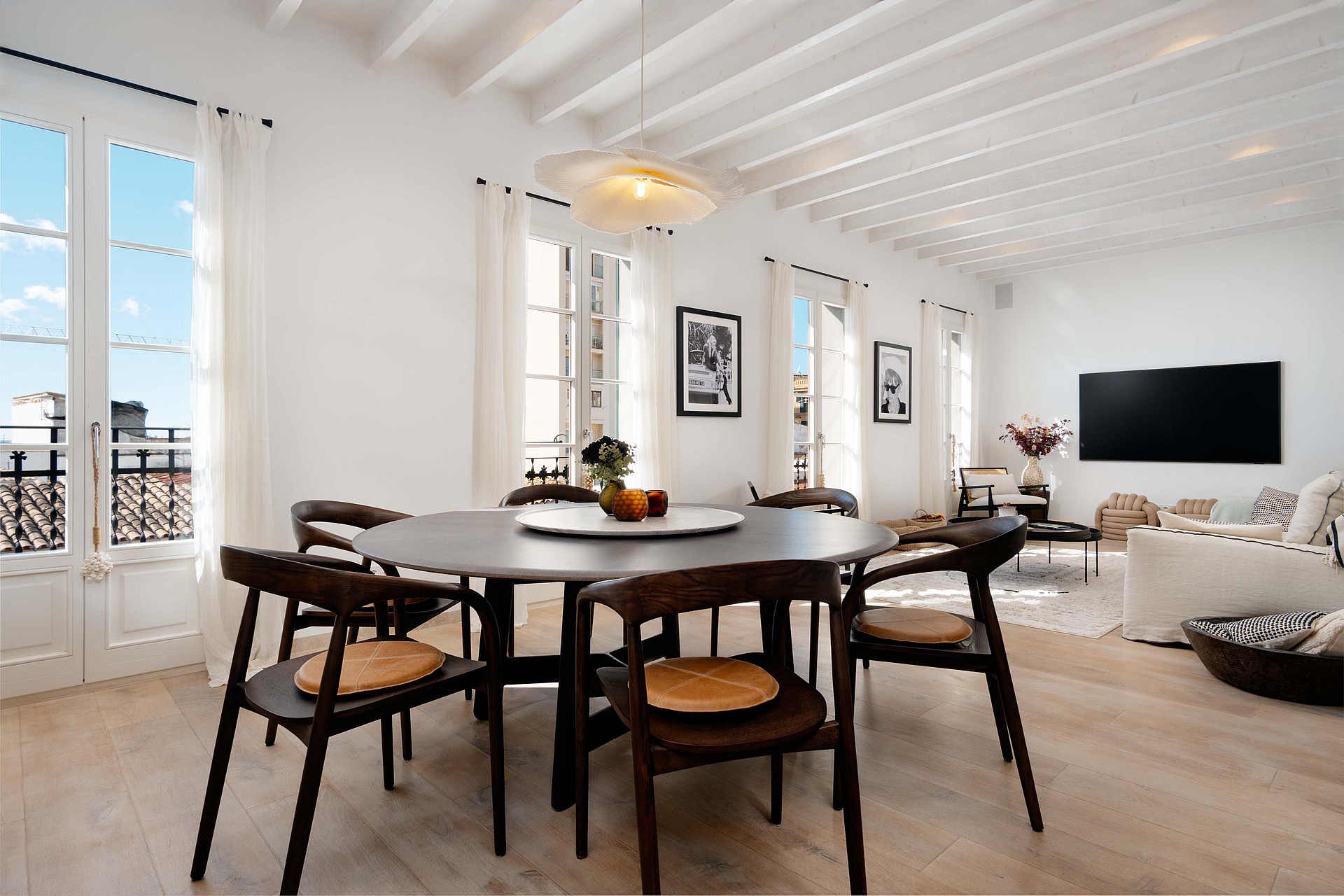 An airy, sunlit open-plan room with a round dark wood dining table, white beamed ceilings, and floor-to-ceiling French doors.